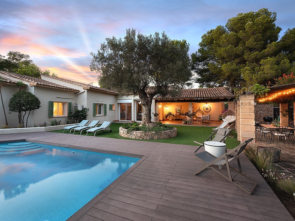 Outdoor swimming pool with wood decking, lounge chairs, and a central olive tree in a villa garden during dusk.