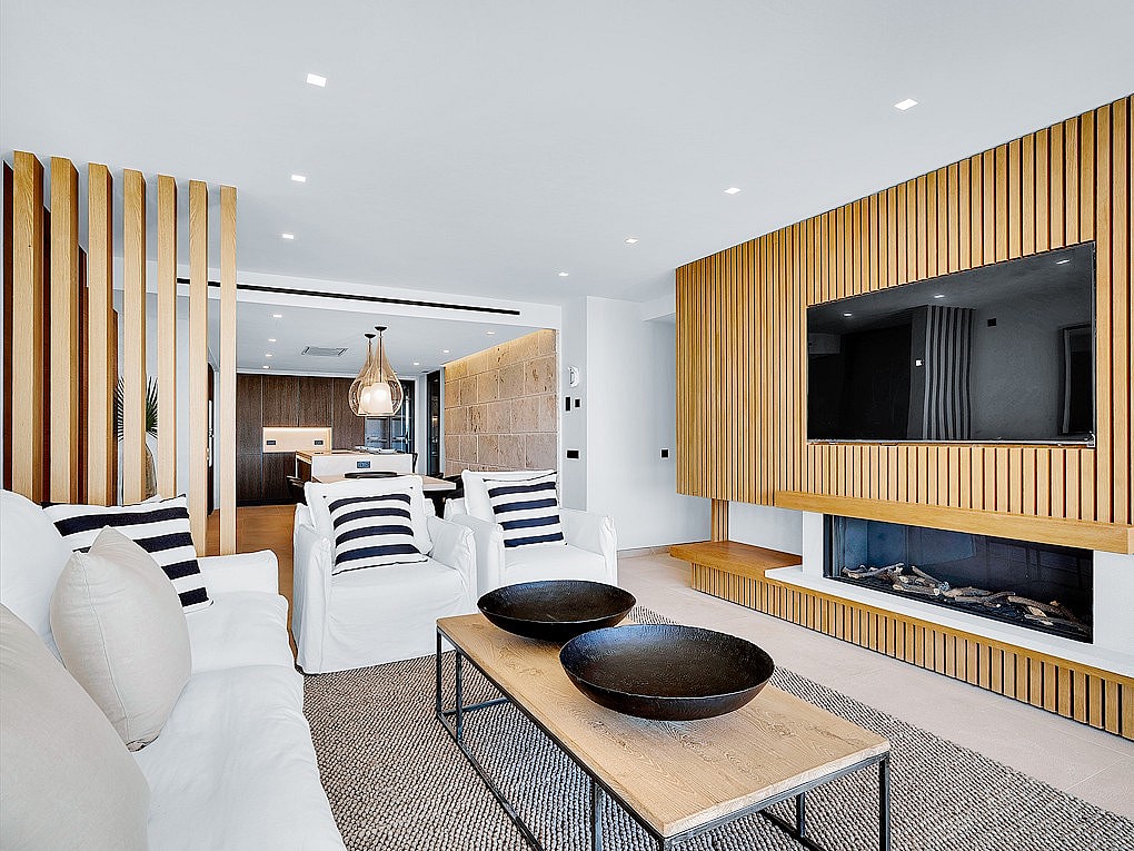 Interior view of a living room featuring a vertical oak slatted wall with an integrated fireplace and TV, white sofas, and coffee table.