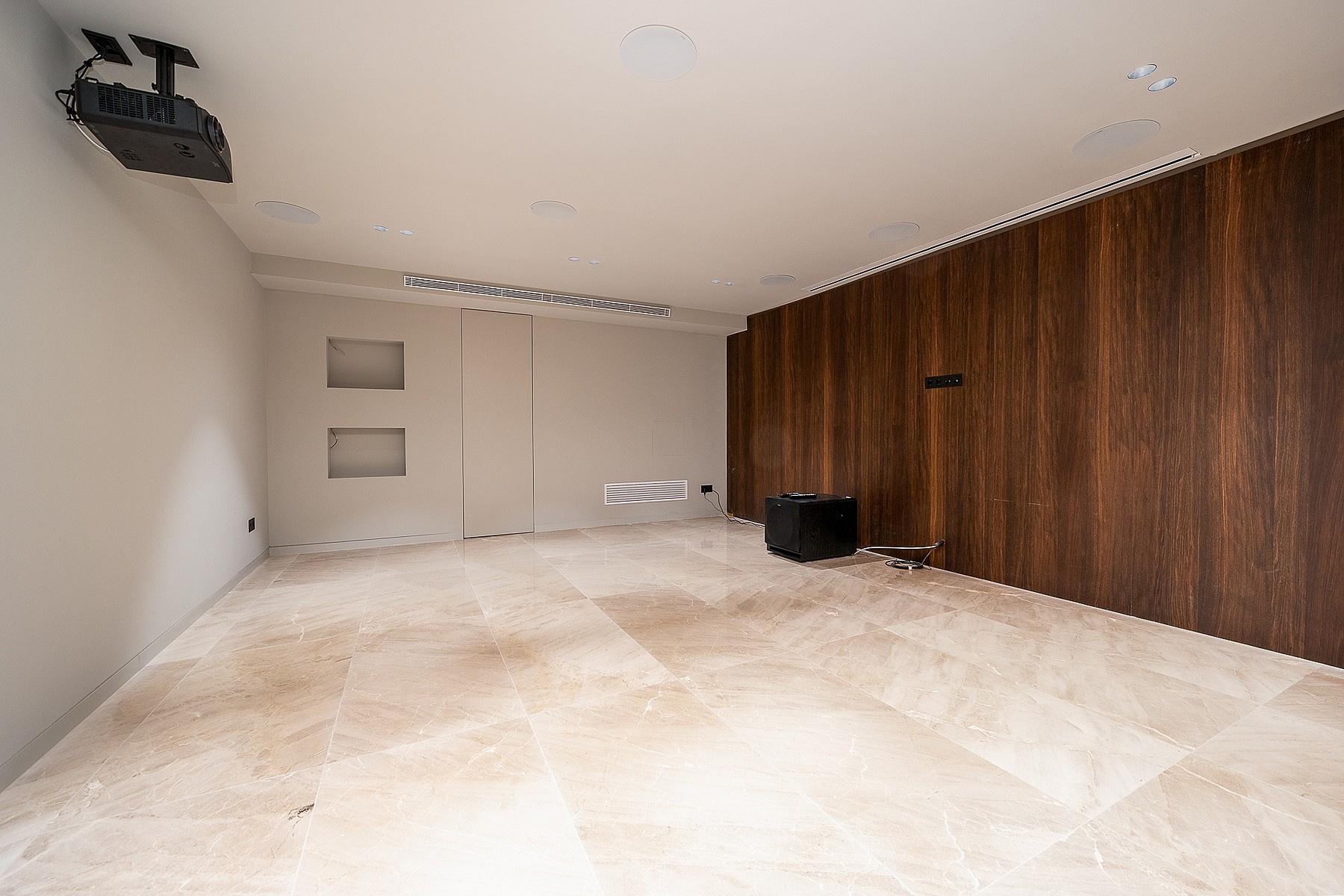 A minimalist home cinema room featuring polished beige stone floors and a dark wood-paneled accent wall, with a ceiling-mounted projector.