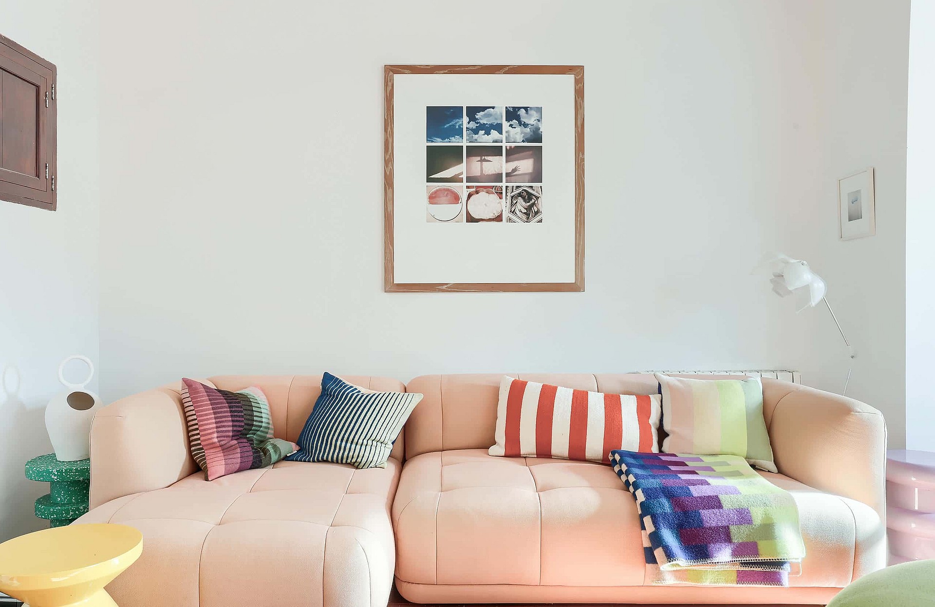 A pale pink sectional sofa sits against a plain white wall, decorated with colorful cushions and a grid-style framed print.