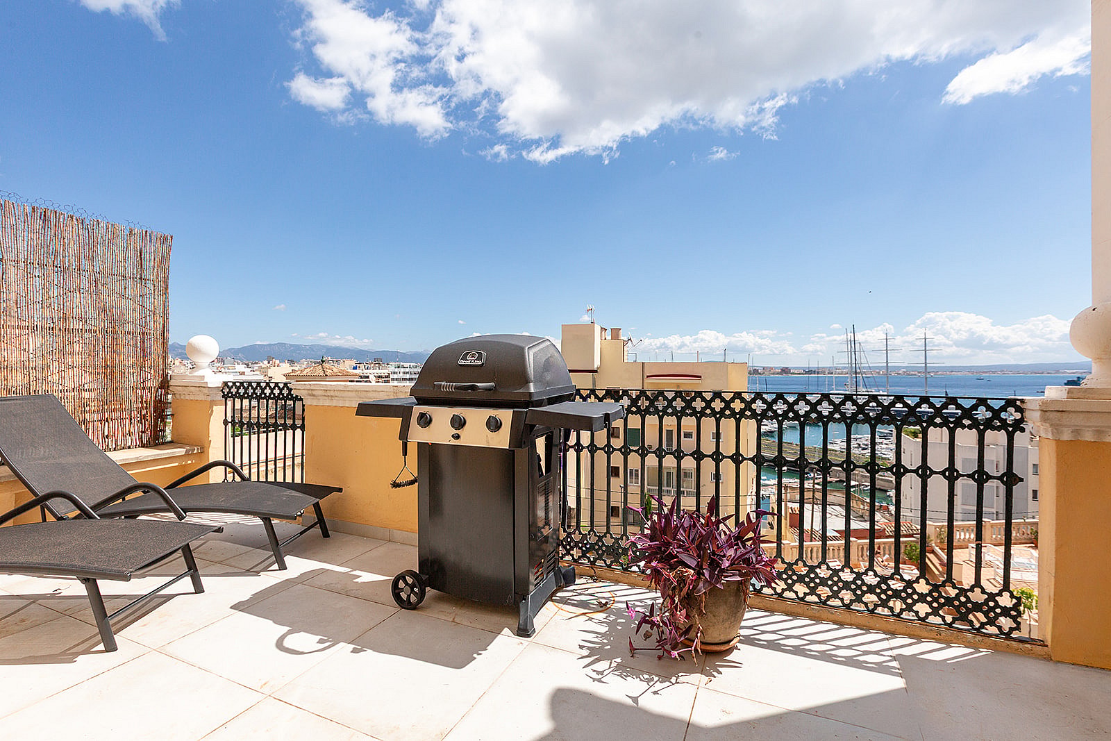 A sunny outdoor terrace featuring two loungers, a portable gas grill, and a wrought-iron railing overlooking the sea.