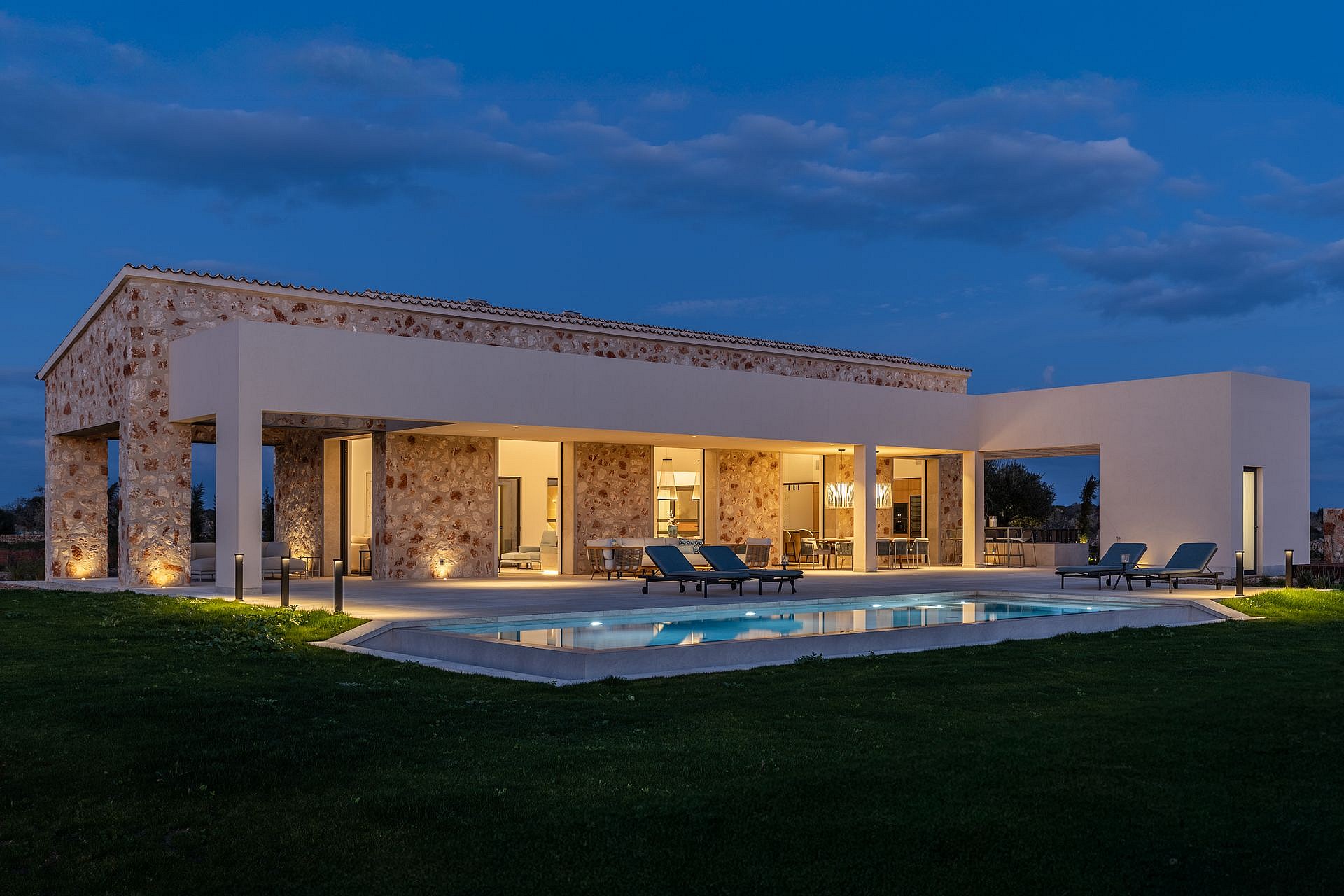 Wide shot of a stone and white plaster finca at dusk, featuring a lit swimming pool and patio furniture on the terrace.