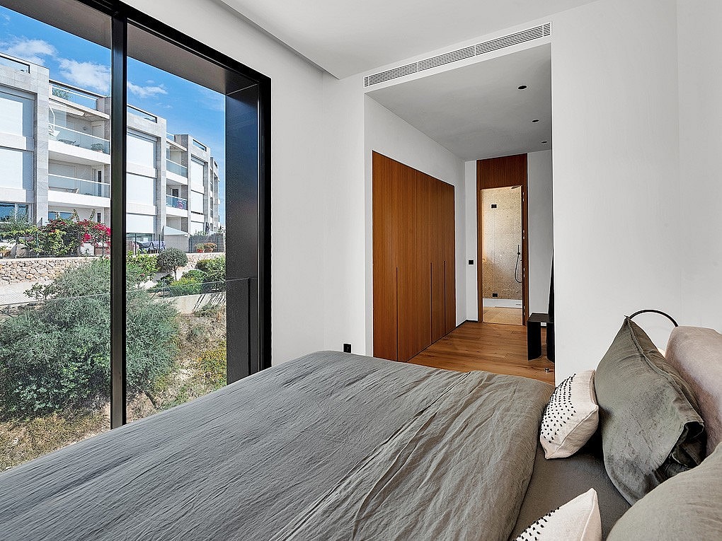 A bedroom featuring floor-to-ceiling windows, a grey textured bed, and wooden wardrobe cabinetry opening to a hallway.
