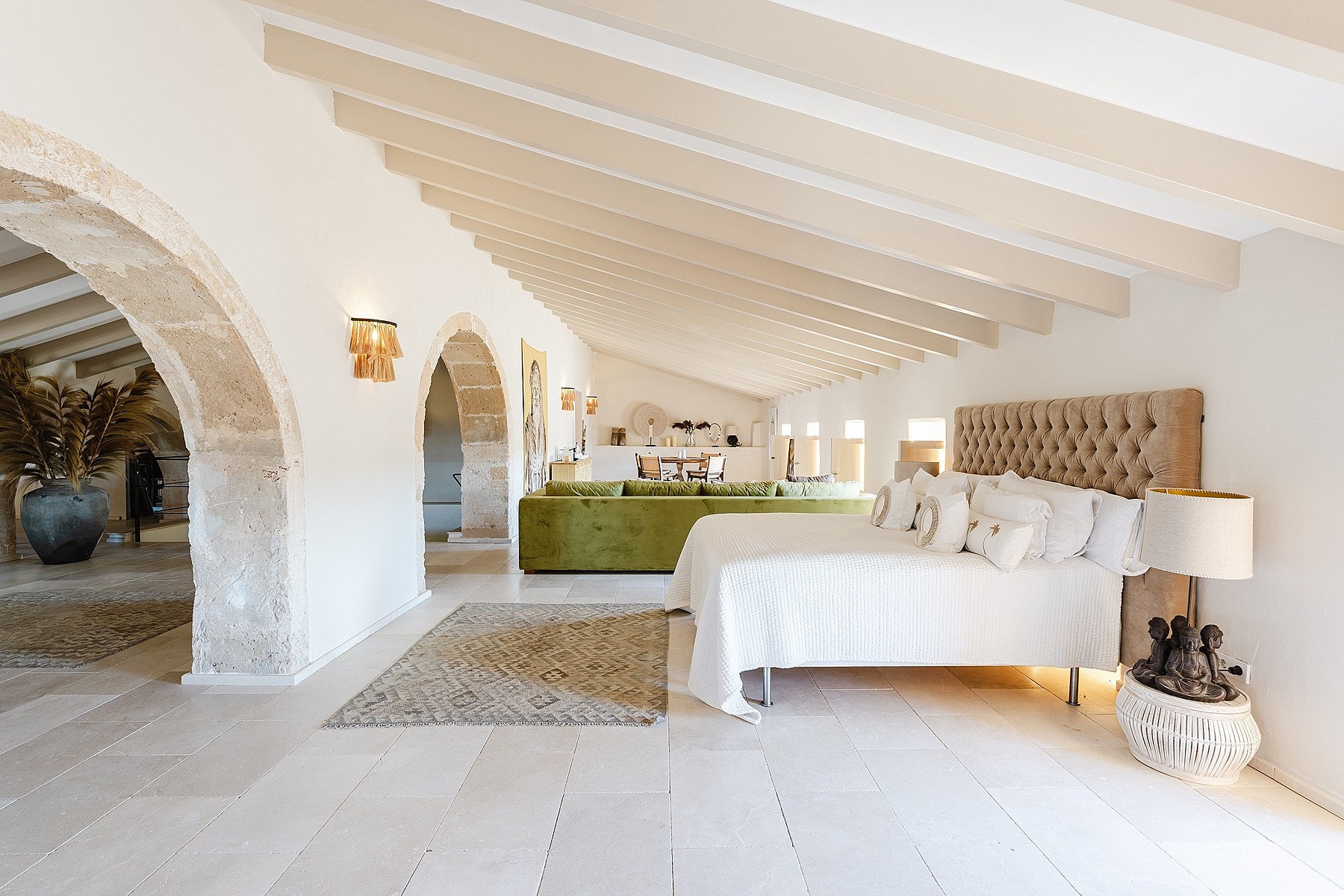 A spacious bedroom with a king-sized bed, tufted headboard, stone flooring, and arched doorways leading to an adjoining area.
