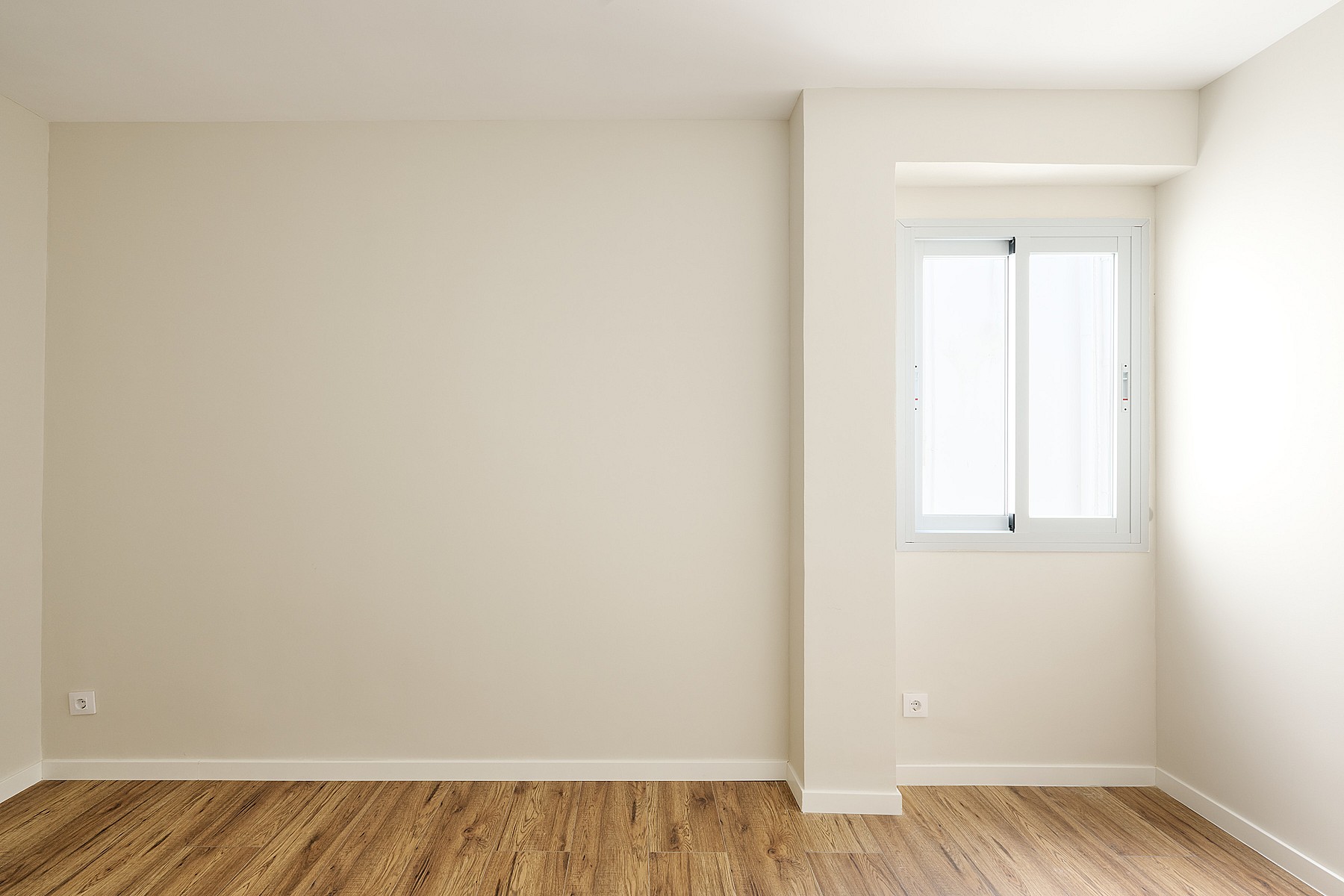 Empty bedroom with light beige walls, wooden floor, and a single white-framed window set into a niche.