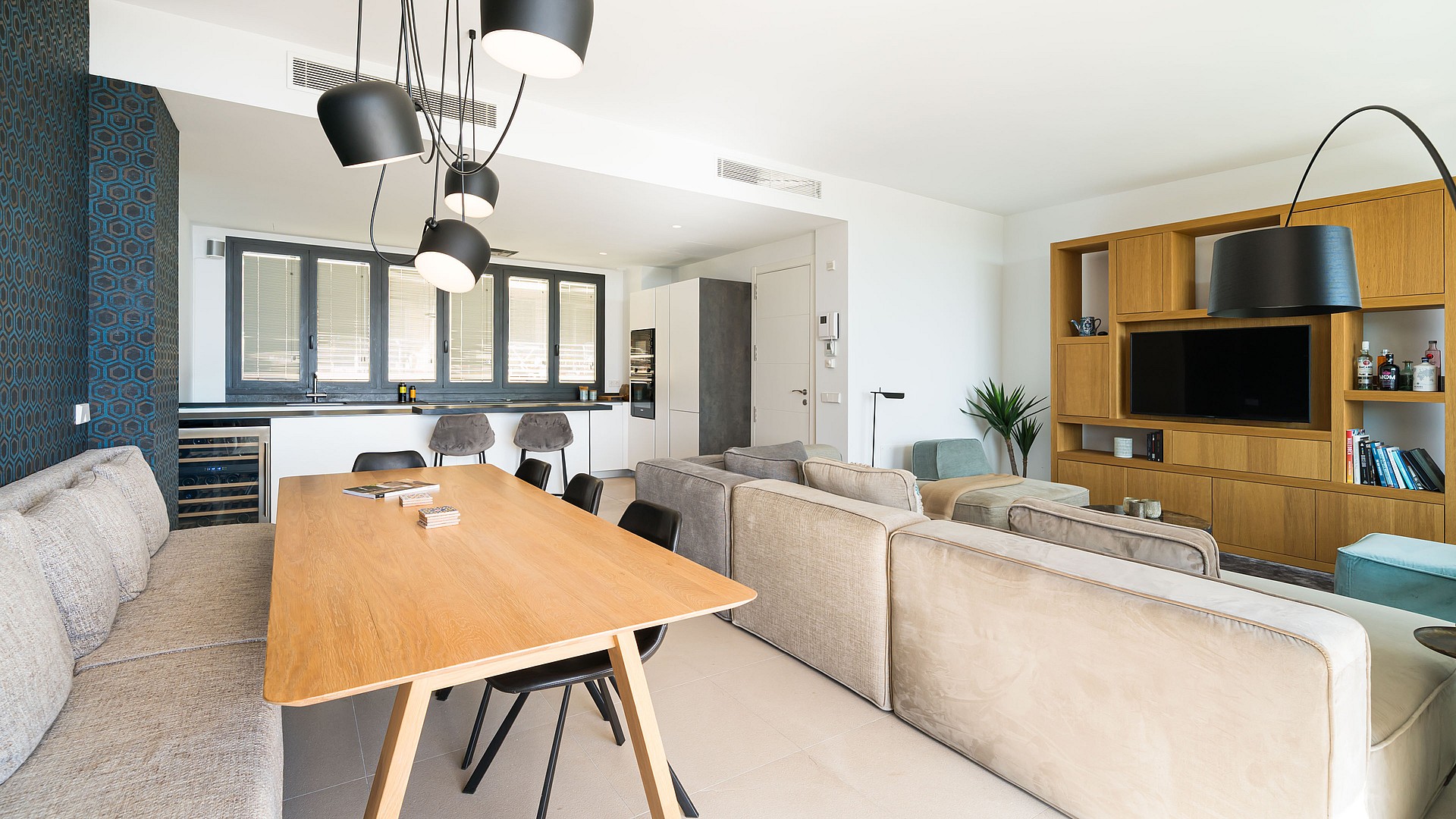 Modern open-plan living room with a beige sectional sofa, light oak wall unit, and a view into a white kitchen.