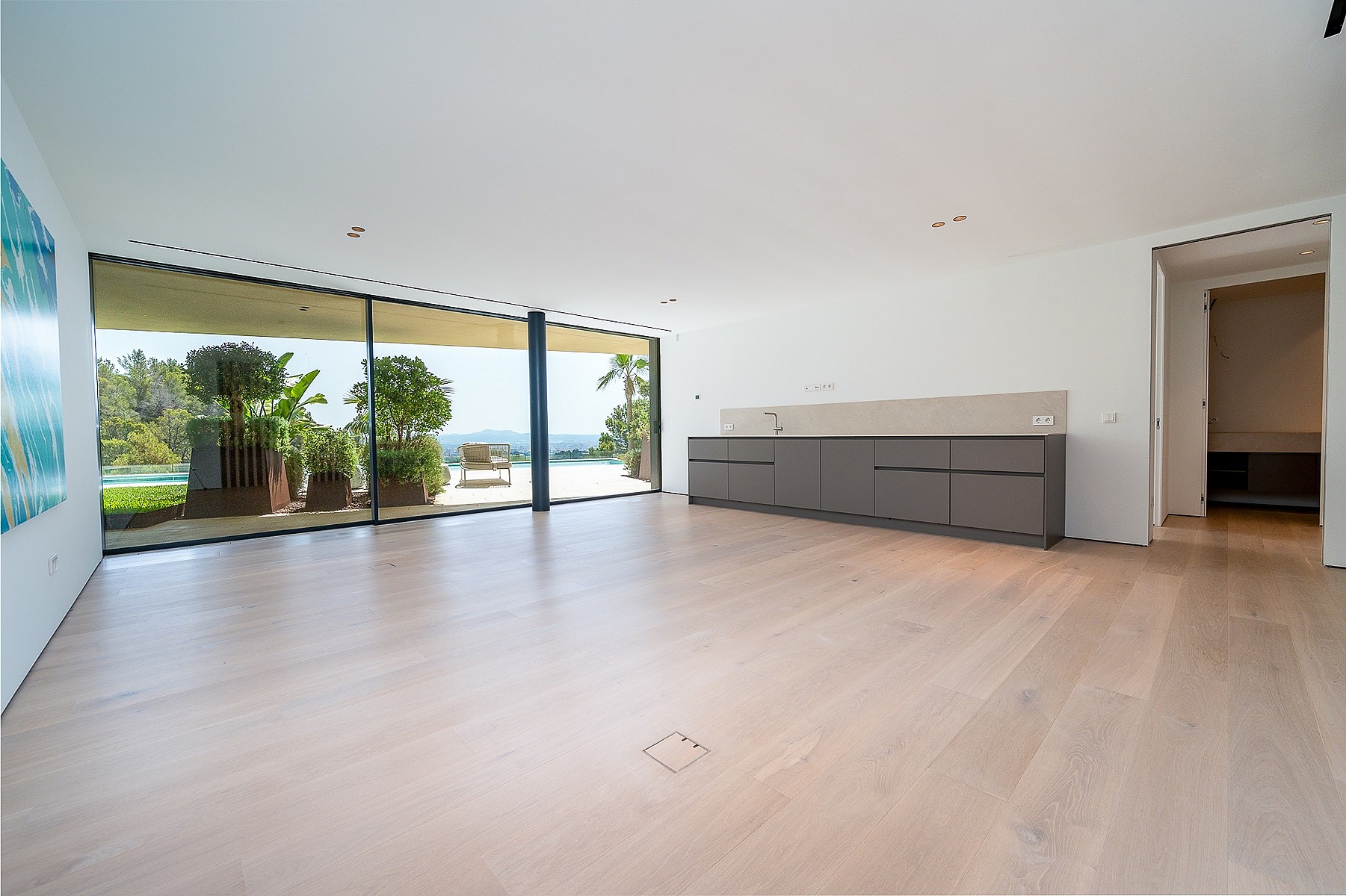 An expansive, unfurnished living room with wide plank light oak flooring, white walls, and a low grey modular cabinet. Floor-to-ceiling glass sliding doors open to a sunlit terrace with greenery and distant sea views.