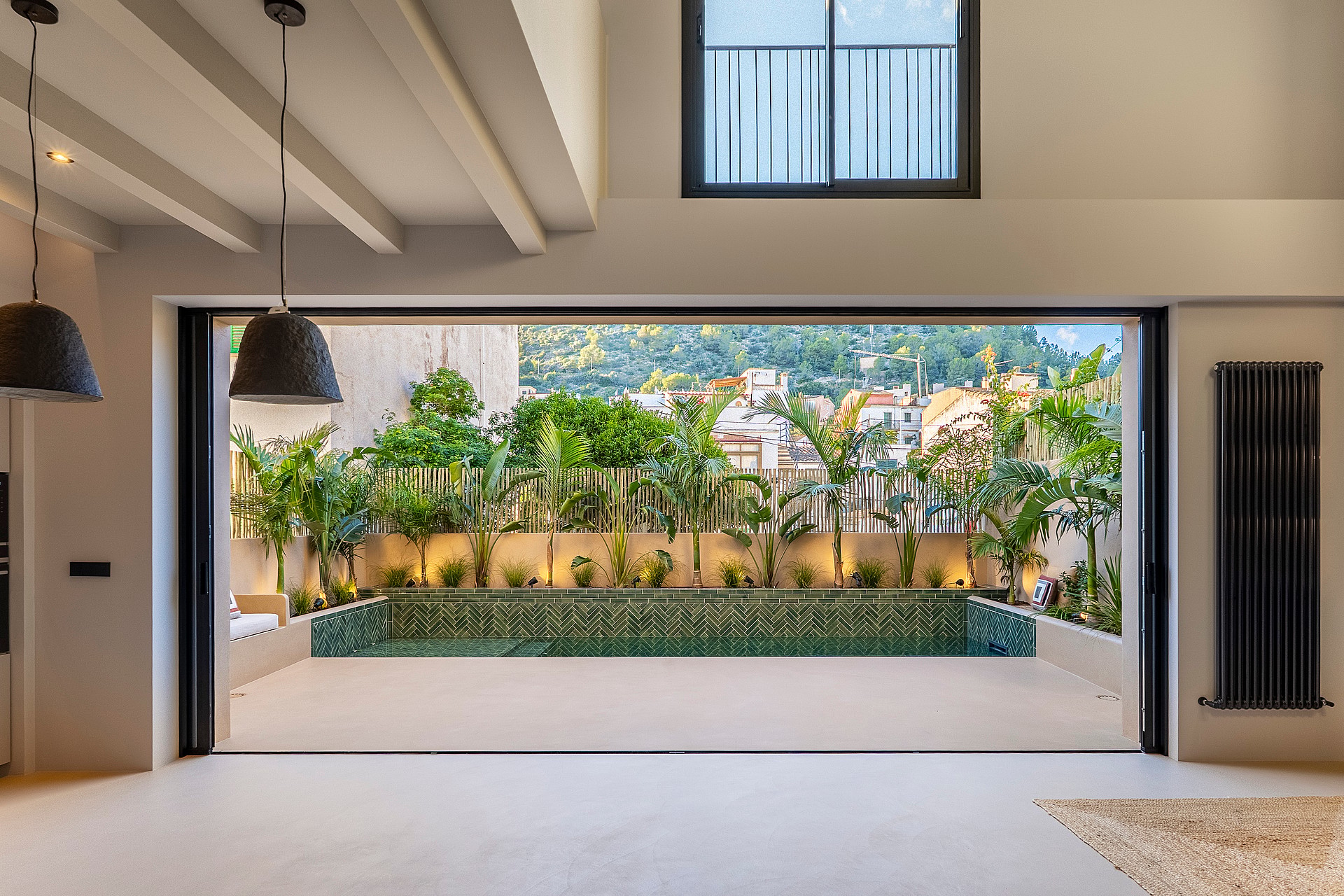 Interior view through open sliding doors to a terrace with a green tiled plunge pool and lush perimeter planting.