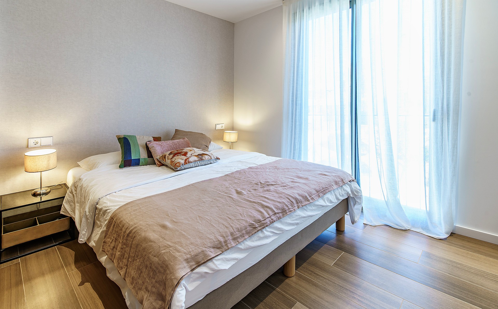 A bright bedroom with a king-sized bed, beige textile headboard, mirrored side tables, and sheer white curtains.