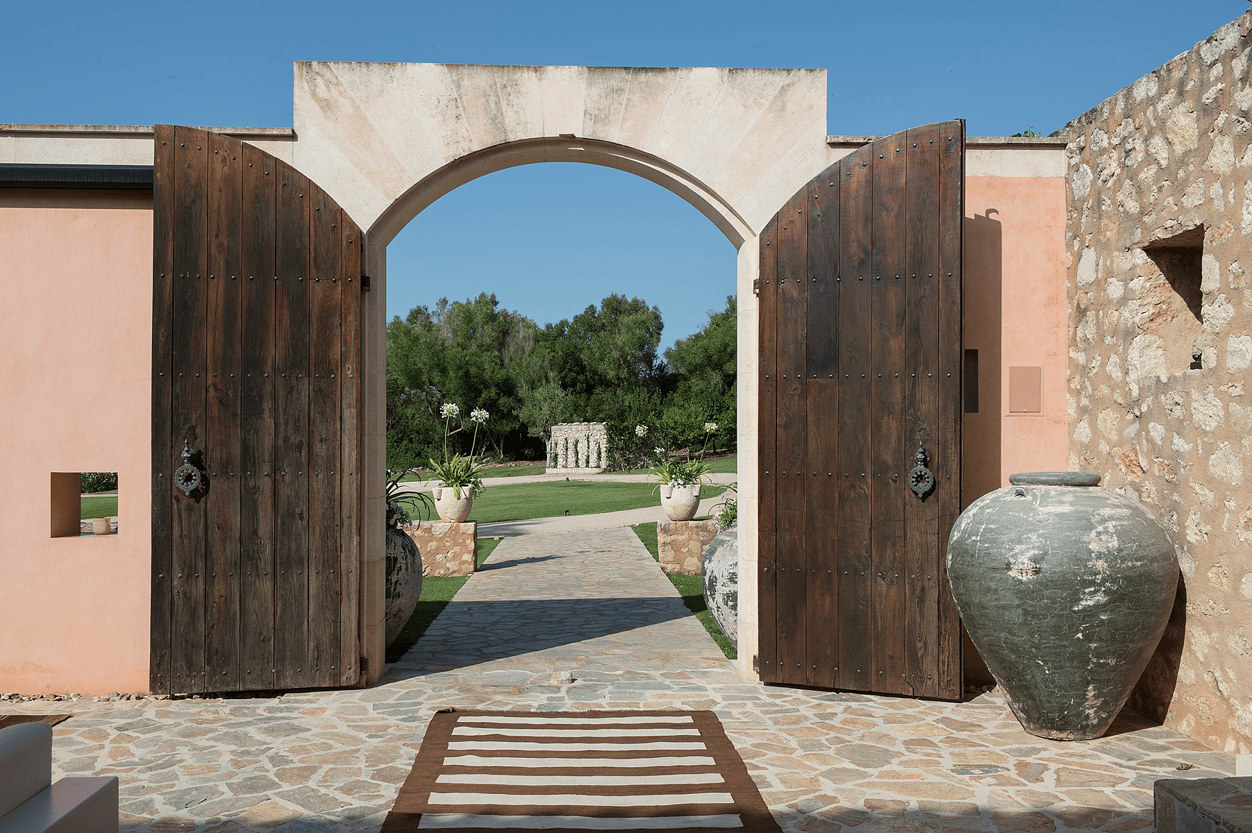 Wide-angle view of an open double-leaf wooden gate leading to a stone-paved courtyard with large terracotta pots.
