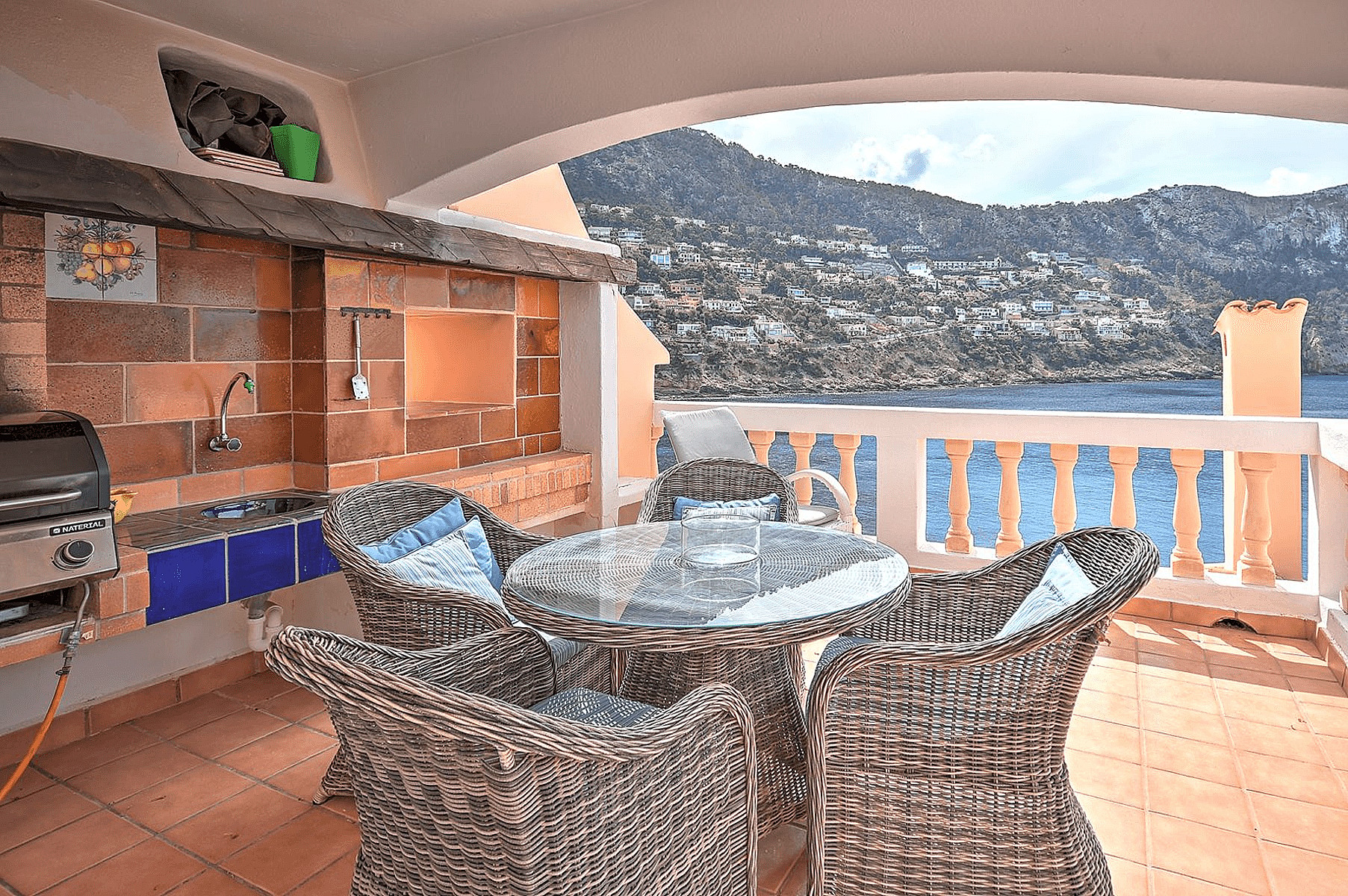Covered terrace featuring an outdoor brick barbecue and kitchen area with dining table and wicker chairs overlooking the sea.