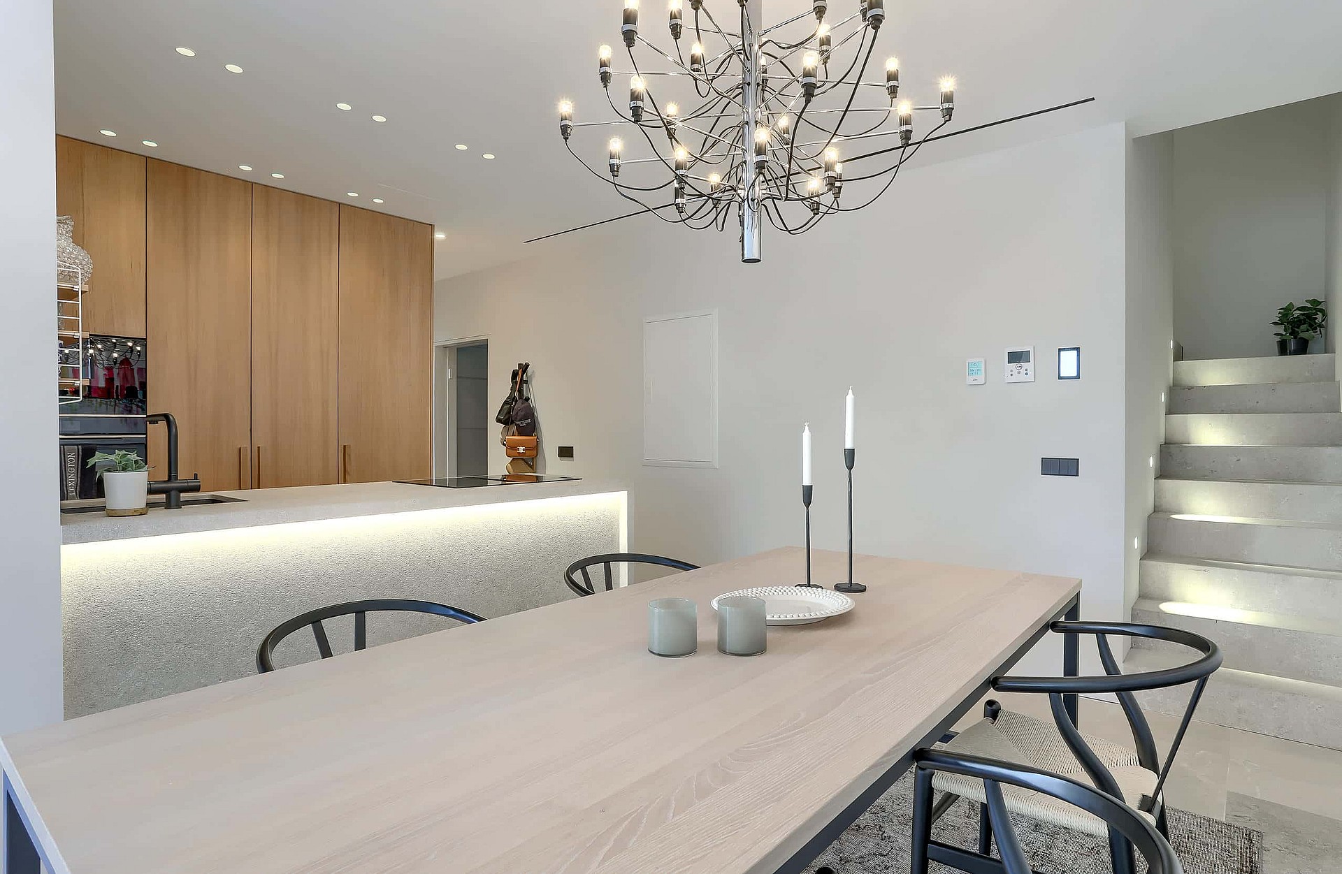 Dining room with light oak table and black chairs beside an open kitchen with illuminated stone island and modern chandelier.