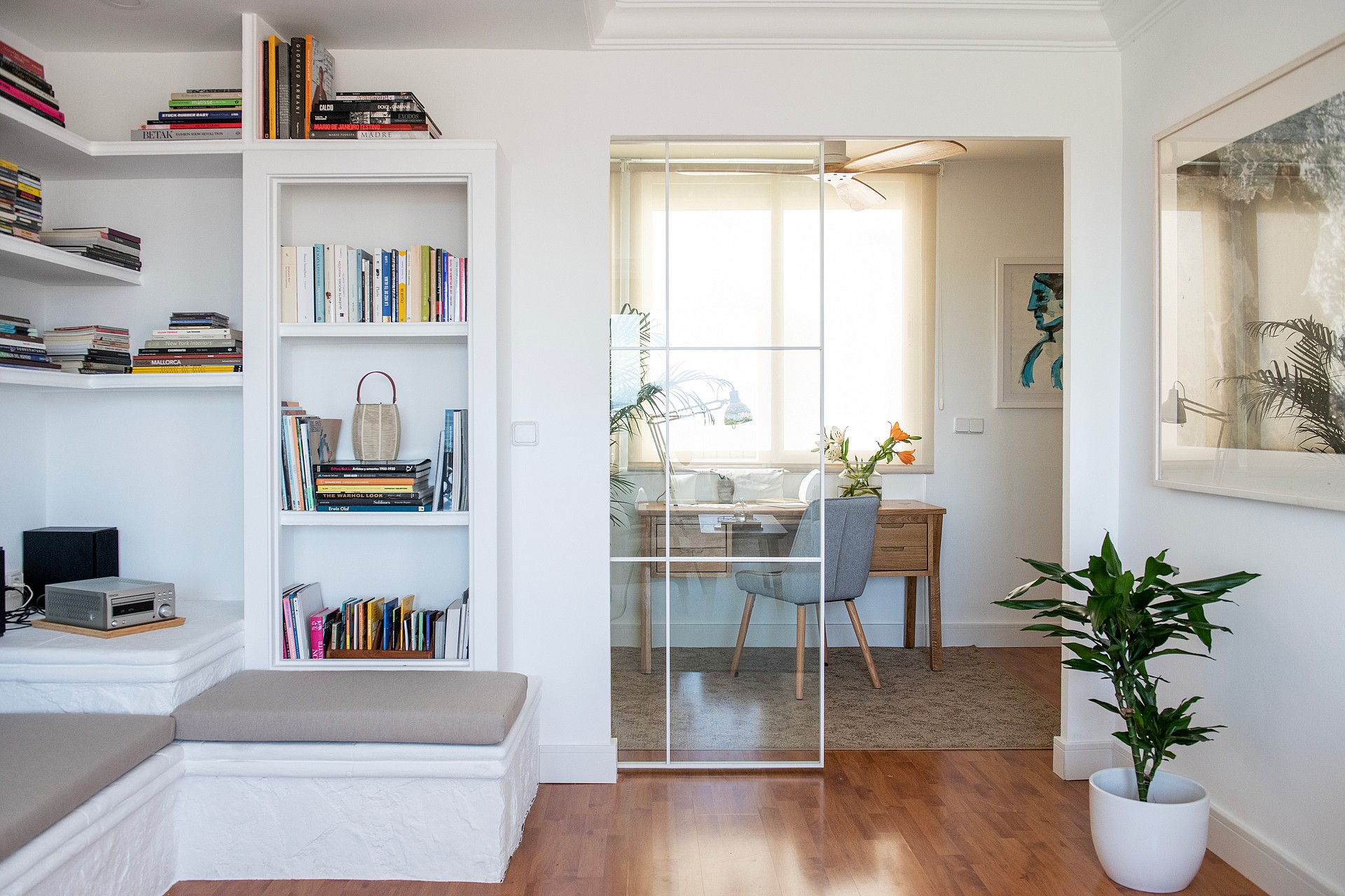 A white built-in bookshelf with a bench seat next to a glass-partitioned office area in a bright penthouse interior.