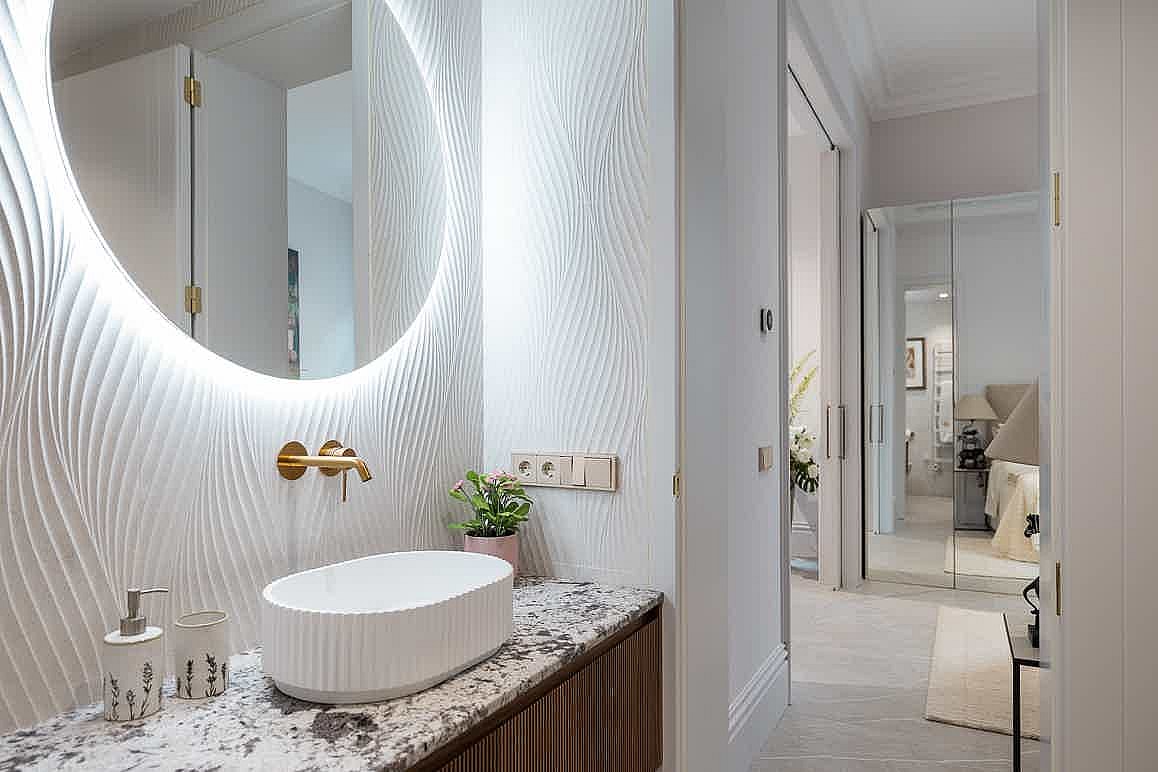 Close-up of a bathroom vanity with a white vessel sink, gold faucet, and a round backlit mirror over a patterned marble wall.