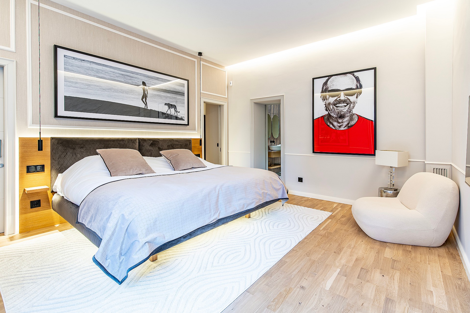Spacious bedroom with a king bed on a light wood floor, soft cream walls, modern pop art, and an integrated white armchair.
