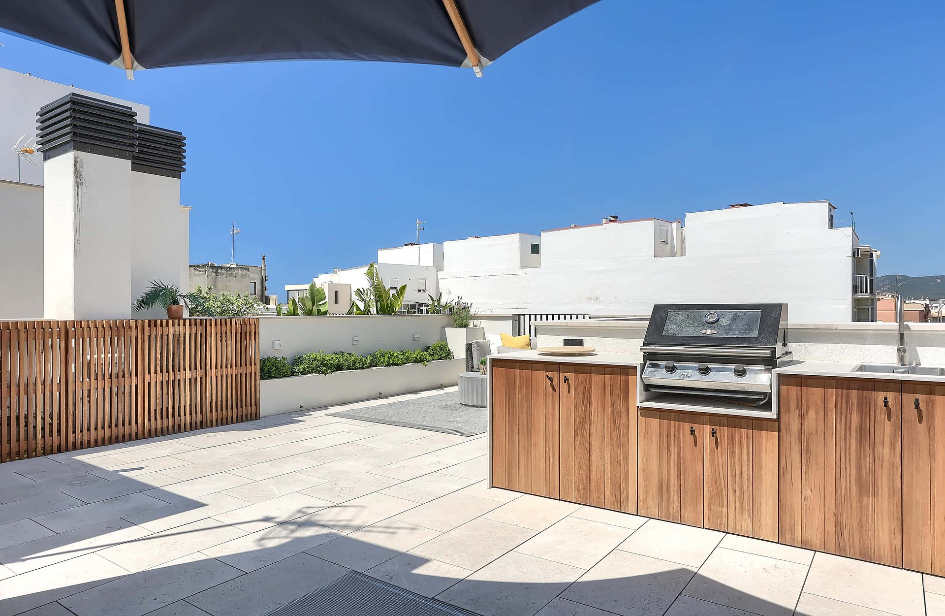 Rooftop terrace featuring a wooden outdoor kitchen cabinet with a built-in stainless steel grill, stone tile flooring, and slatted wood privacy fencing under a clear blue sky.