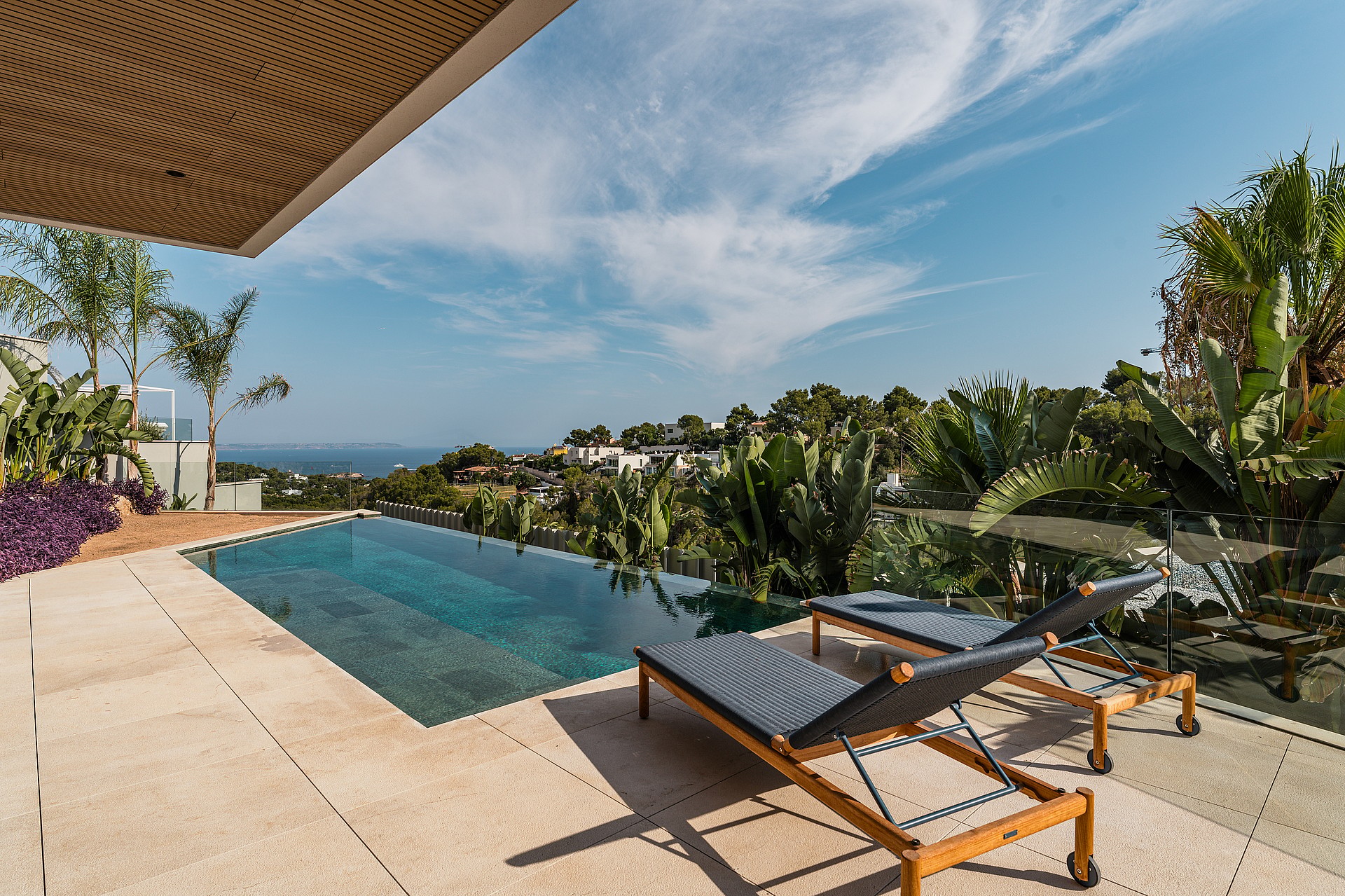 Two dark wicker sun loungers with wooden frames on a stone terrace overlooking an infinity pool and Mediterranean foliage.