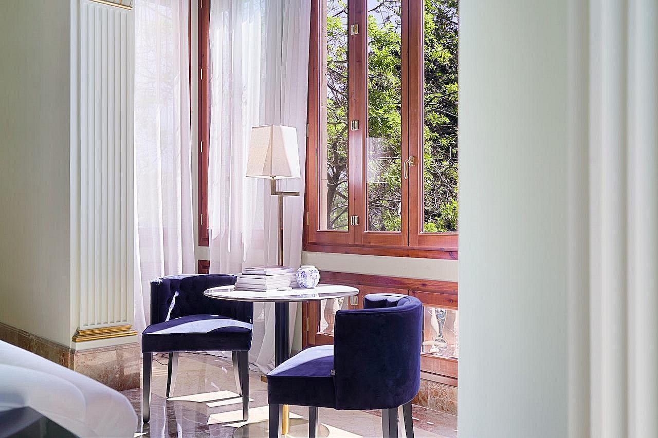 A small circular table with two blue upholstered chairs placed beside tall wood-framed windows with white curtains.