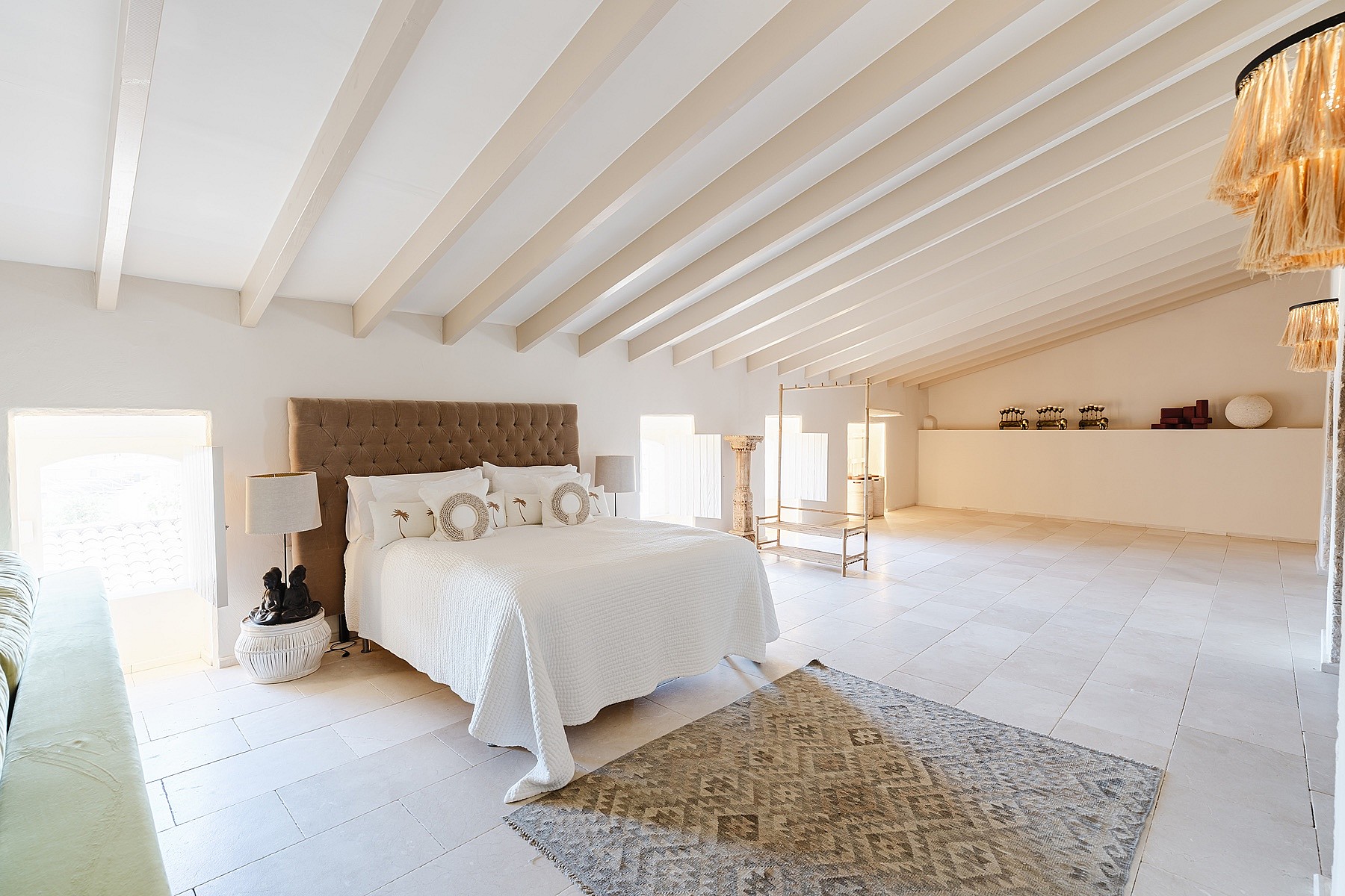 Spacious bedroom with a tufted headboard, white linens, and light wood rafters in a bright, minimalist attic space.