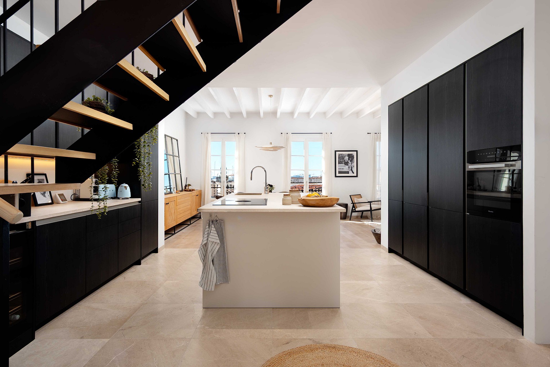 An open-plan kitchen in a Santa Catalina apartment featuring black cabinetry, a white central island, and a dark metal staircase.