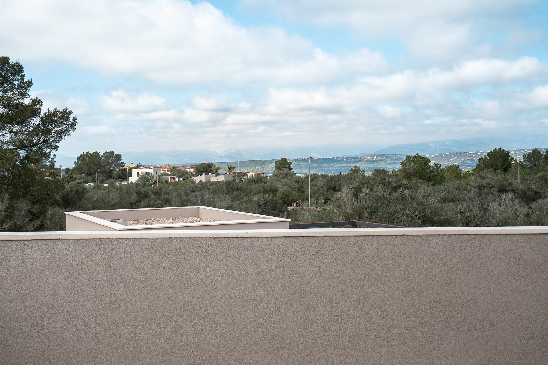 A view from a terrace overlooking a green landscape of olive trees with distant mountains under a cloudy sky.