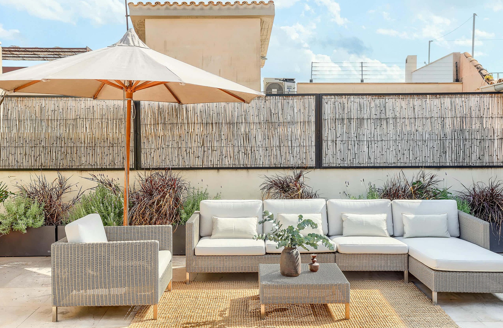 A rooftop terrace featuring a modular wicker sofa set with white cushions, a matching coffee table, and a large beige parasol, set against a backdrop of Old Town buildings.