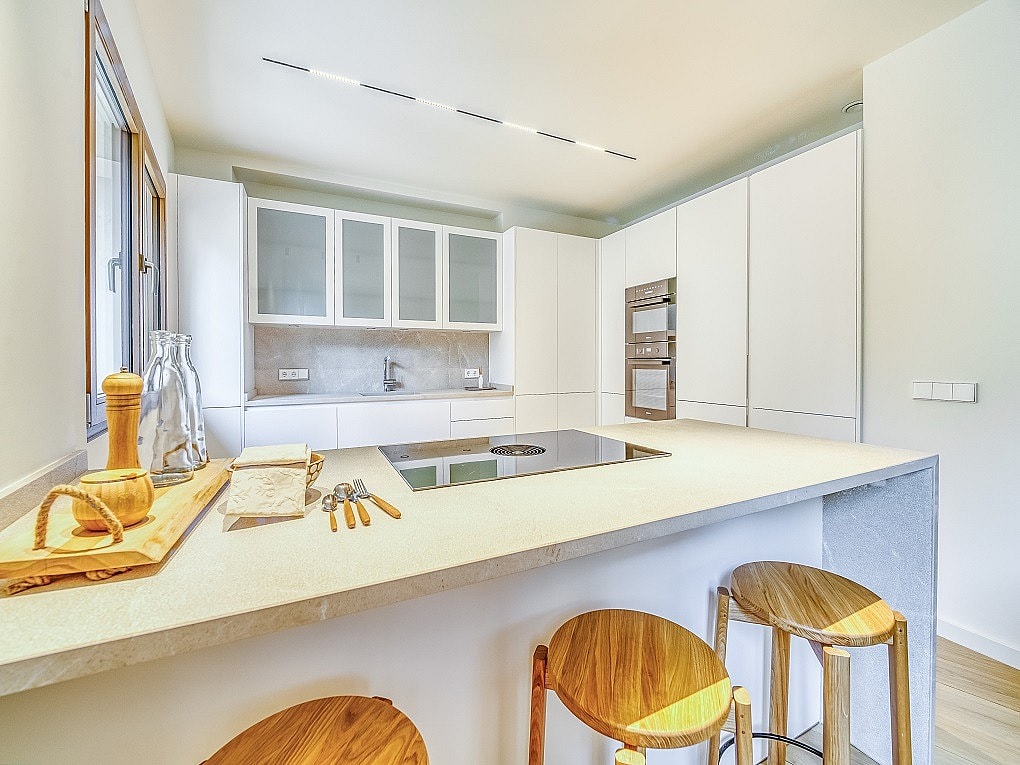 Modern kitchen with white cabinetry, integrated appliances, and a light stone island with wooden bar stools in the foreground.