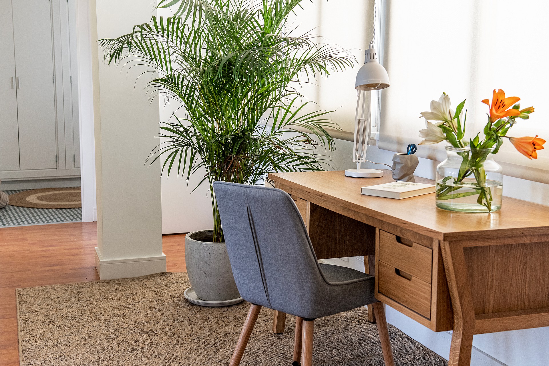 A wooden desk with a grey upholstered chair and a tall potted plant in a bright, light-filled corner of the penthouse.