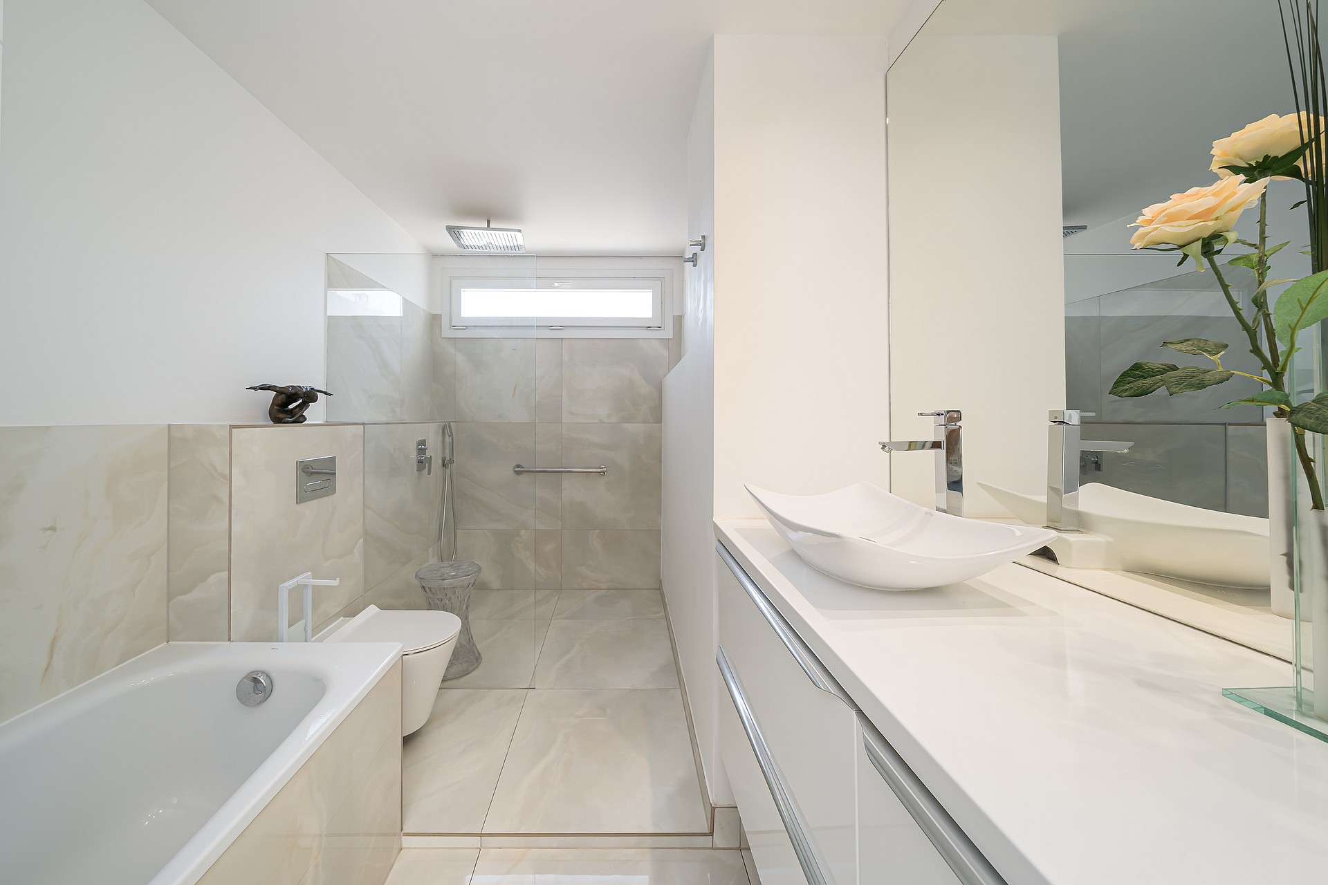 Interior of a modern bathroom with large light-toned floor tiles, a wall-hung toilet, a bathtub, and a sleek vanity sink.