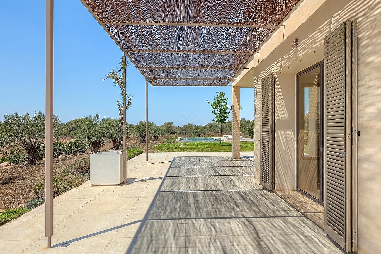 A wide outdoor terrace featuring a woven reed pergola casting dappled shadows on grey stone flooring, next to the finca wall.