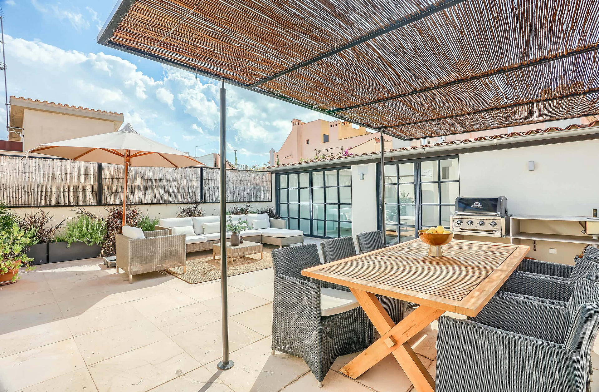 A spacious outdoor terrace with a wooden dining table, wicker chairs, and a shaded lounge area against a city backdrop.