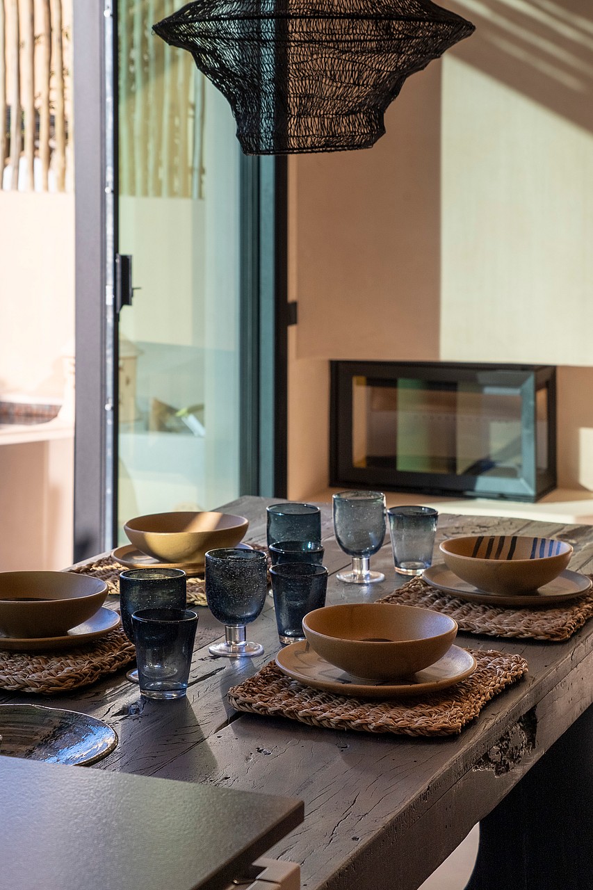 A close-up view of a dining table set with rustic ceramic bowls, blue glassware, and woven placemats, featuring a textured pendant lamp above.