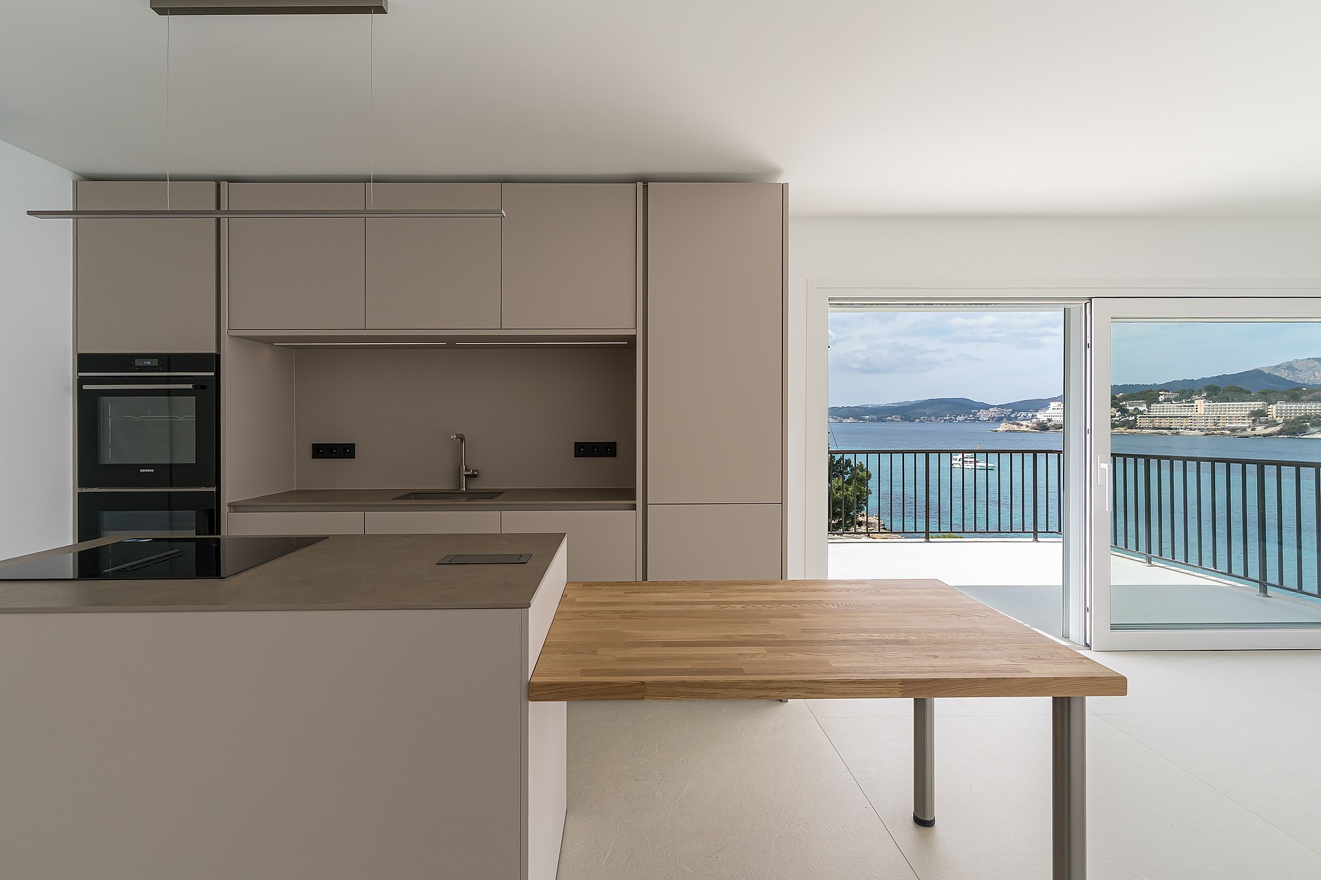 Minimalist kitchen with taupe cabinetry and an oak dining surface opening onto a sea-view terrace.