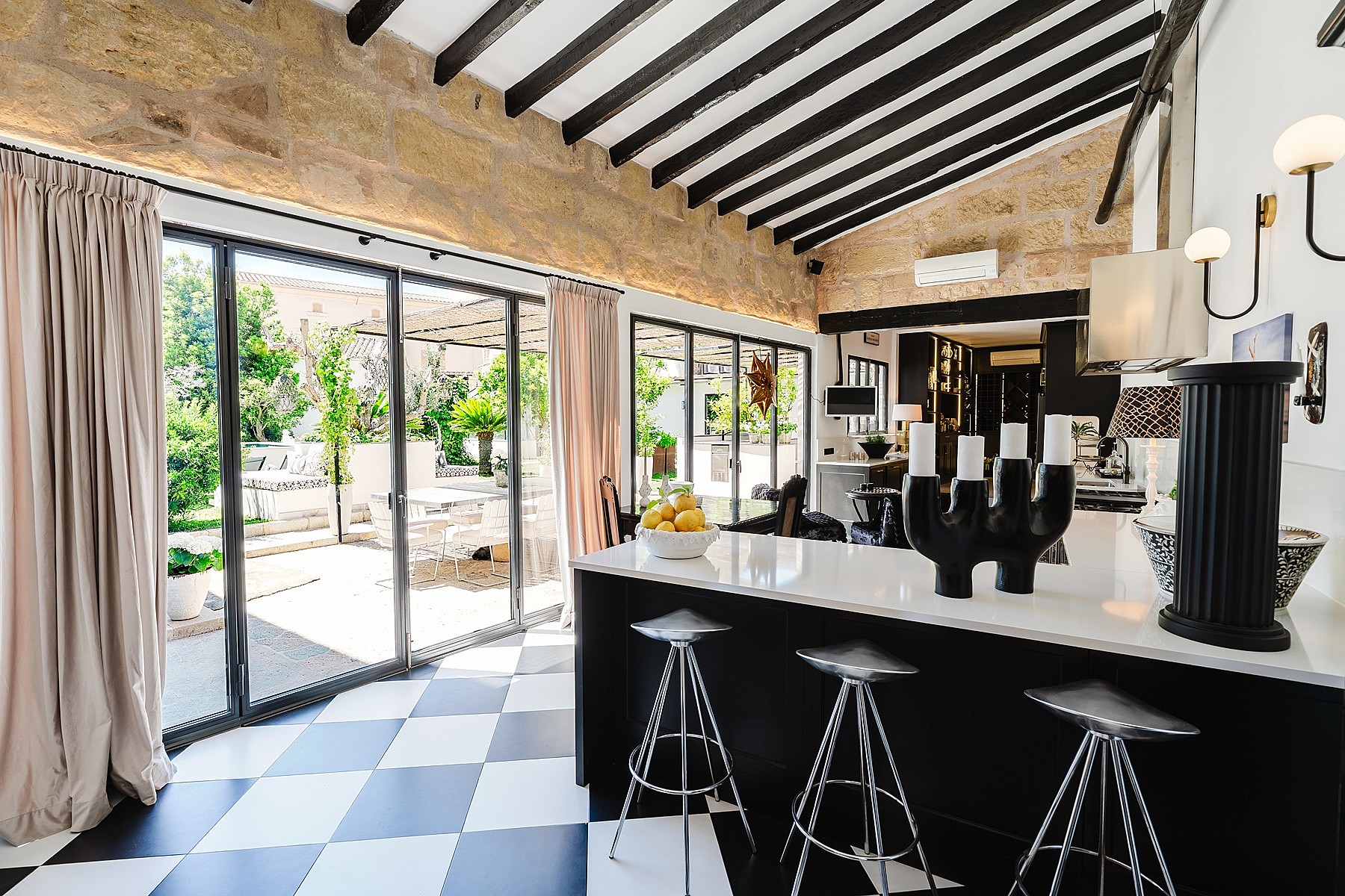 Modern kitchen with black cabinetry, white countertops, checkered floors, and exposed stone walls under a high beamed ceiling.