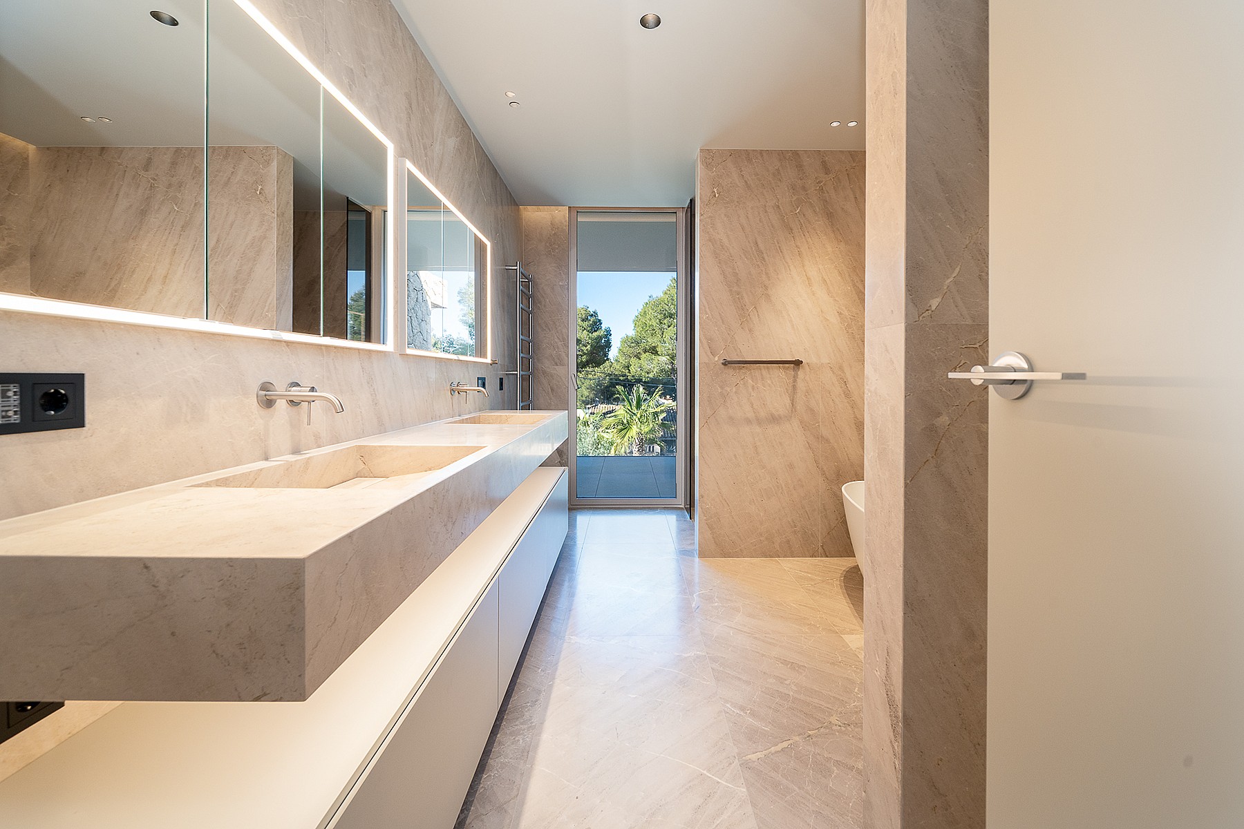 Modern bathroom featuring a long, wall-mounted stone double vanity, integrated LED lighting, and a glass-paneled garden view.