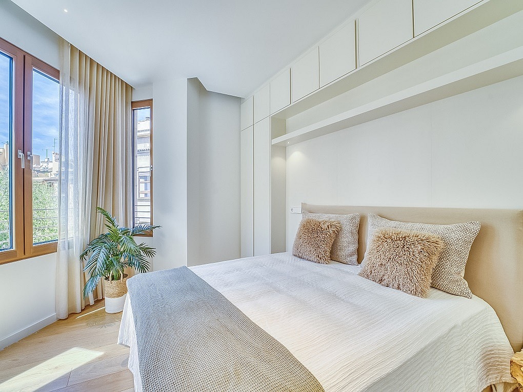 A bright bedroom with a double bed, integrated ceiling-height white wardrobes, and windows showing an urban view.