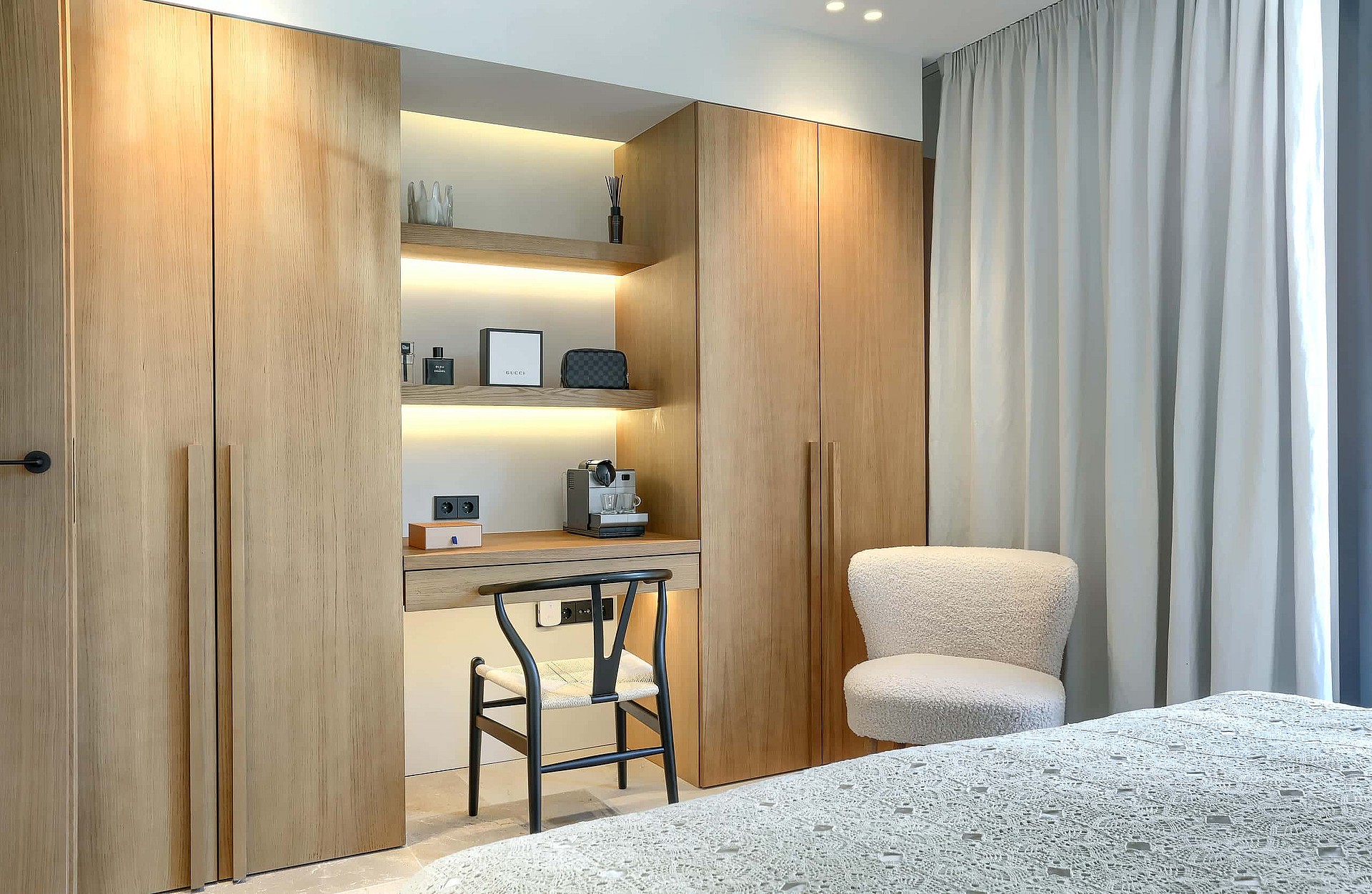 A modern bedroom featuring a floor-to-ceiling light oak wardrobe unit with a built-in desk, shelving, and a black wishbone chair.