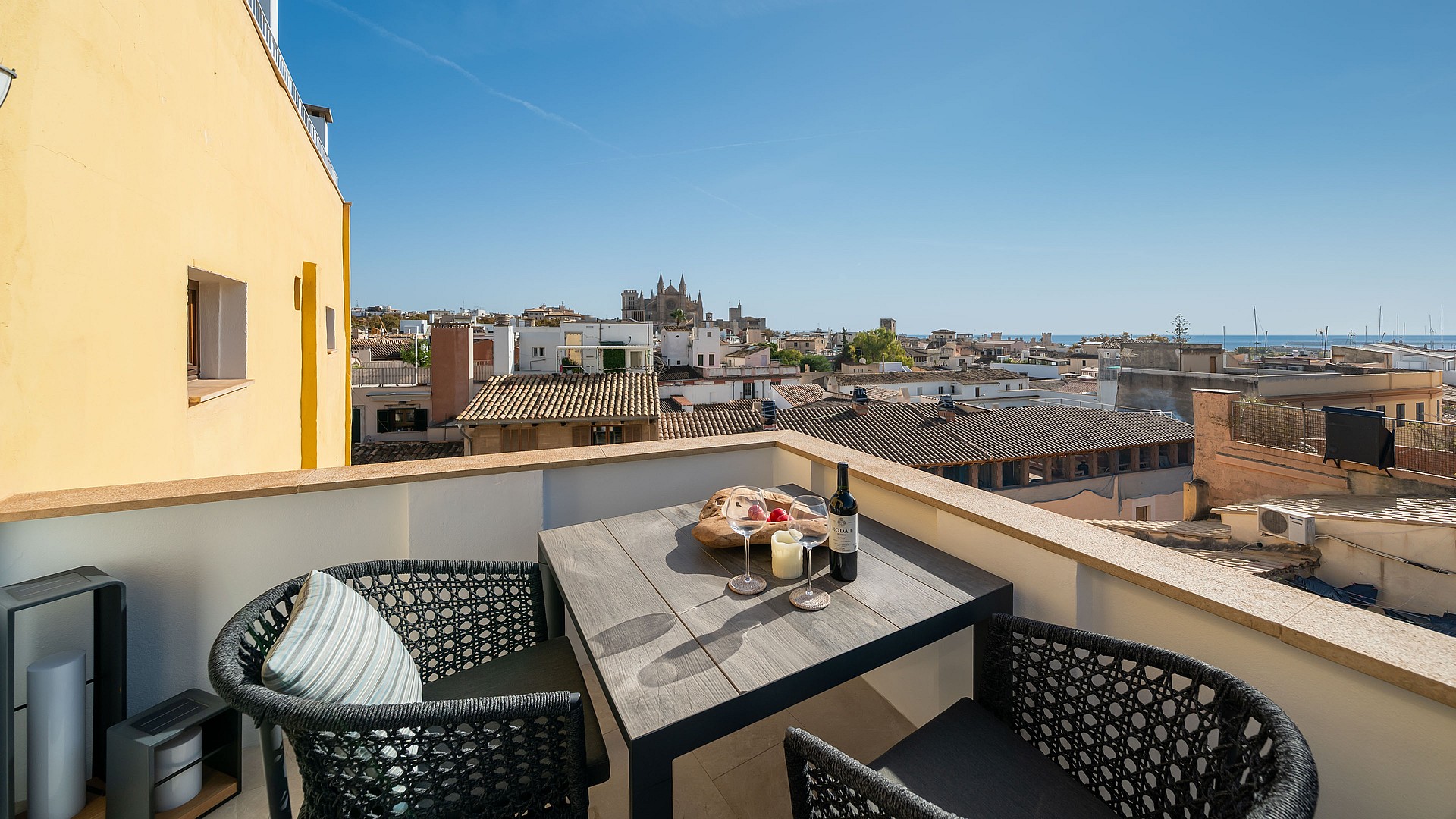 Rooftop terrace with wooden dining table and woven chairs overlooking historic Palma rooftops and the cathedral.