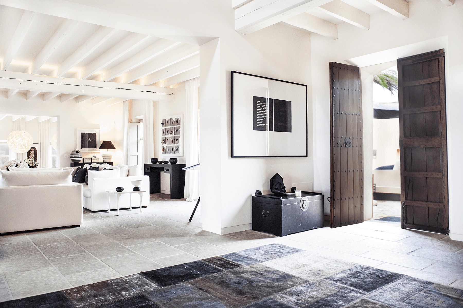 A bright living room with white walls, exposed wooden ceiling beams, stone flooring, and dark wood doors opening to the outside.