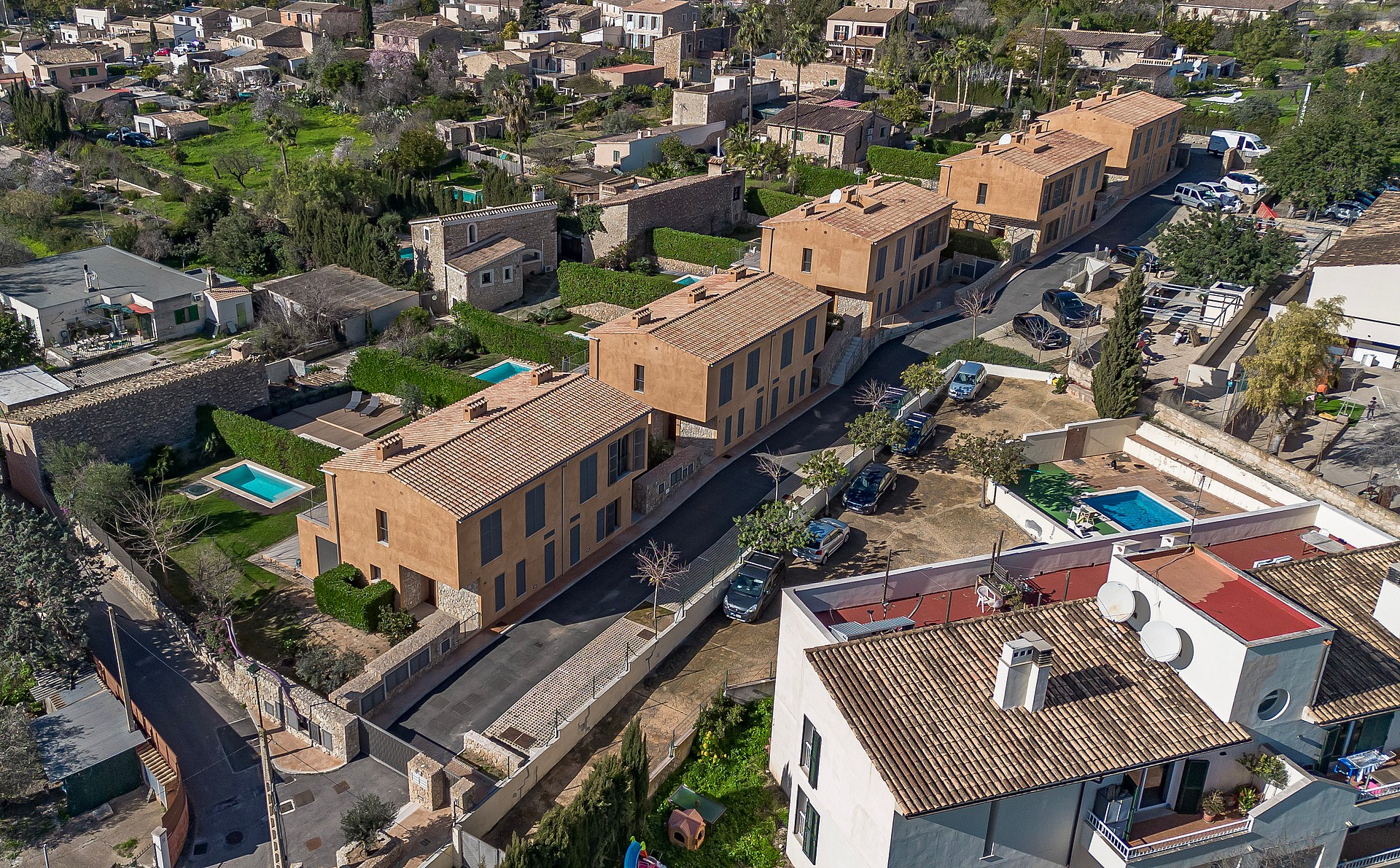Aerial drone shot showing a row of stone-clad townhouses with terracotta tile roofs, private gardens, and small pools.