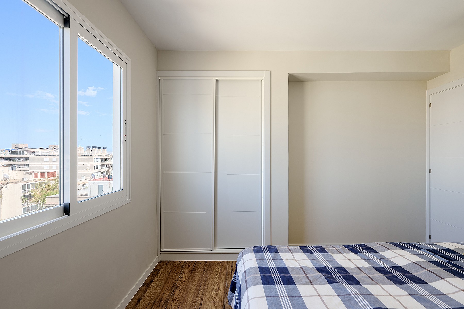 A minimalist bedroom featuring light walls, a wooden floor, and built-in sliding wardrobes, with a window view of city buildings.