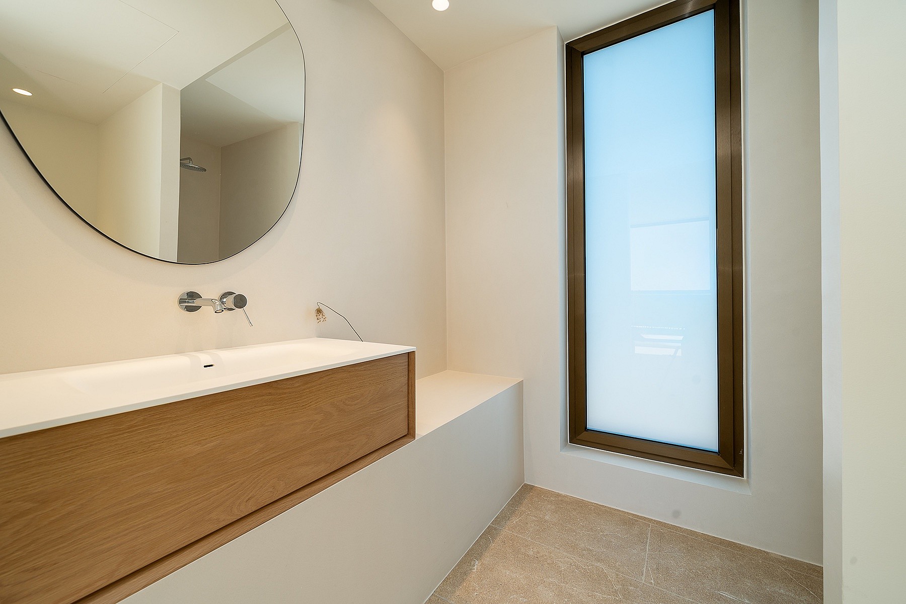 Modern bathroom with a wall-mounted oak vanity, circular mirror, and tall frosted window allowing diffused natural light.