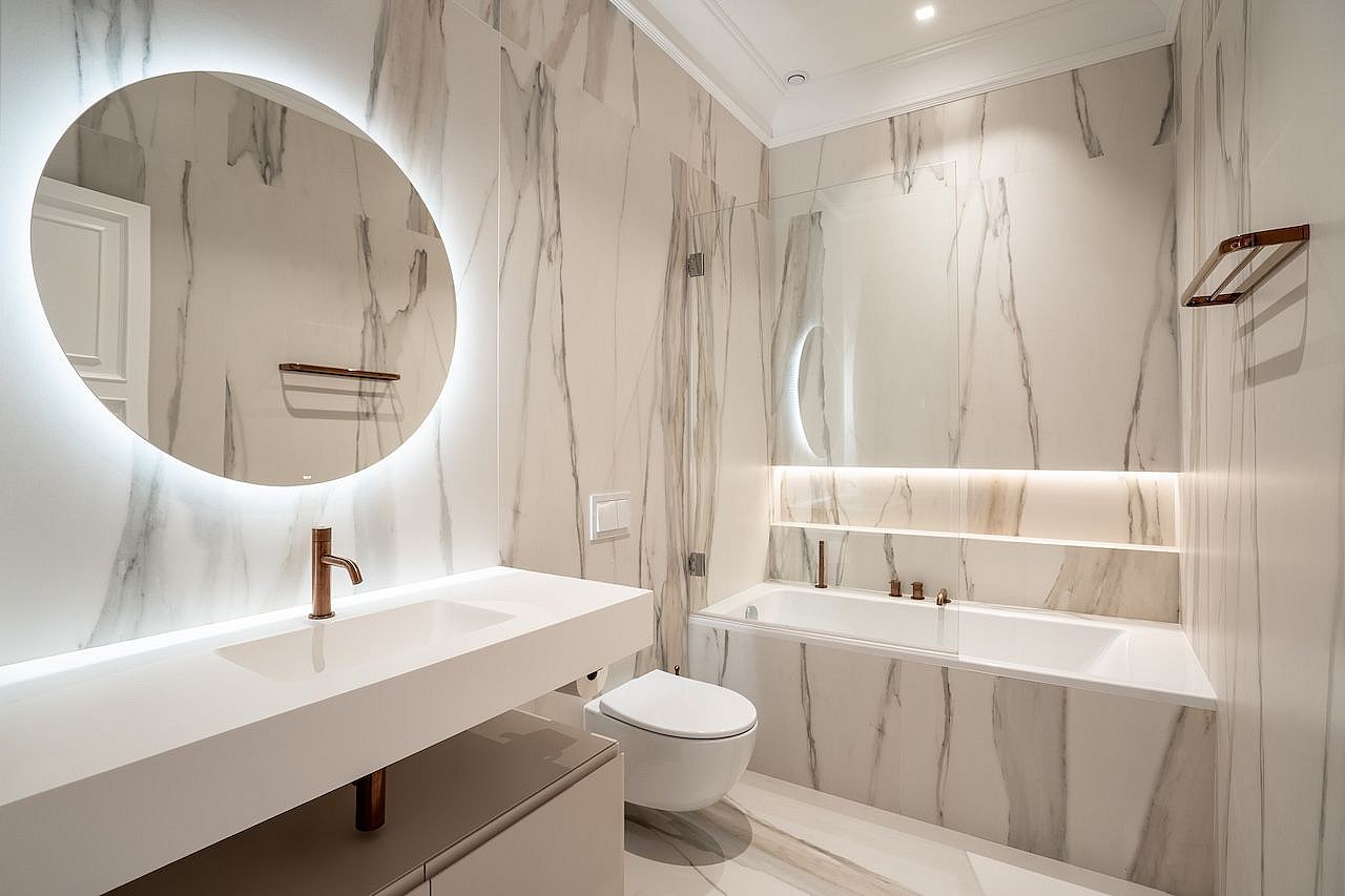 A modern bathroom featuring floor-to-ceiling white marble with grey veining, a wall-mounted vanity, and a backlit circular mirror.