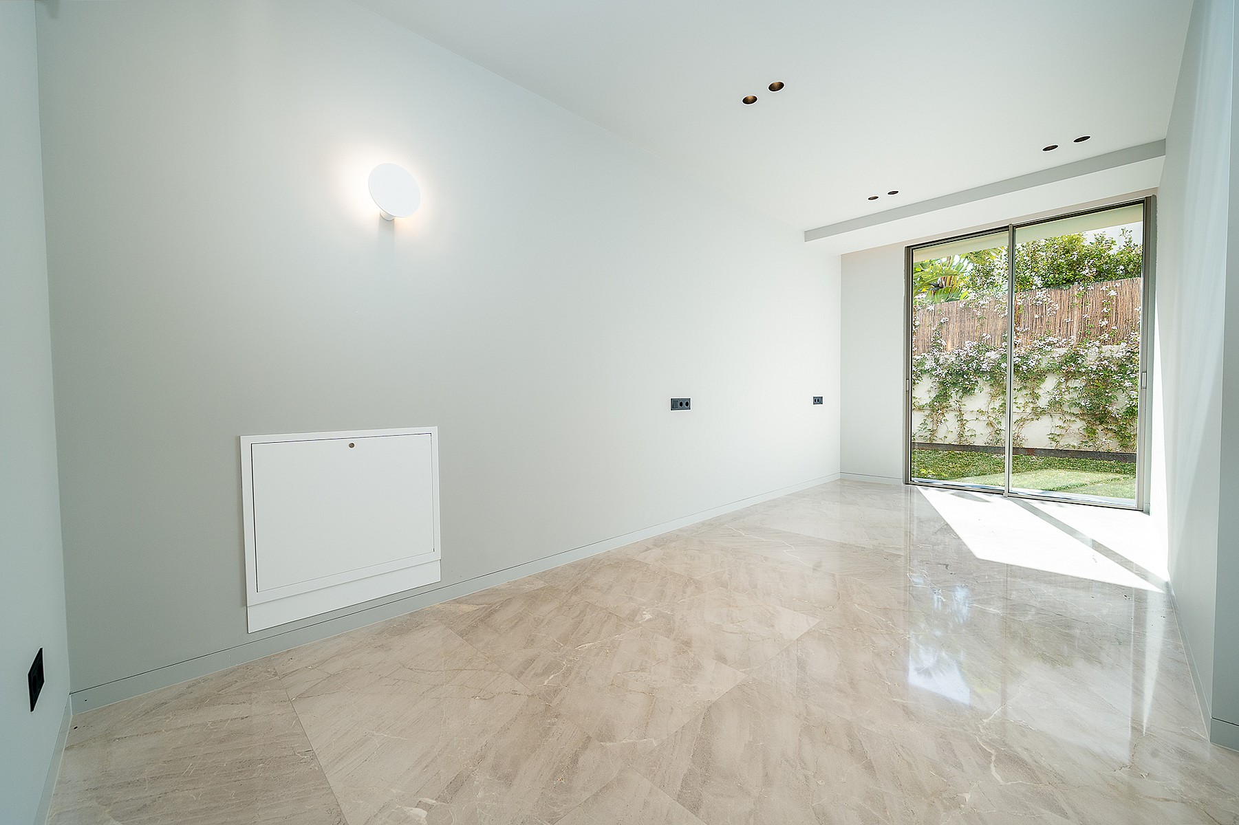 Empty rectangular room with polished marble floors, plain white walls, and a large sliding glass door opening to a garden.