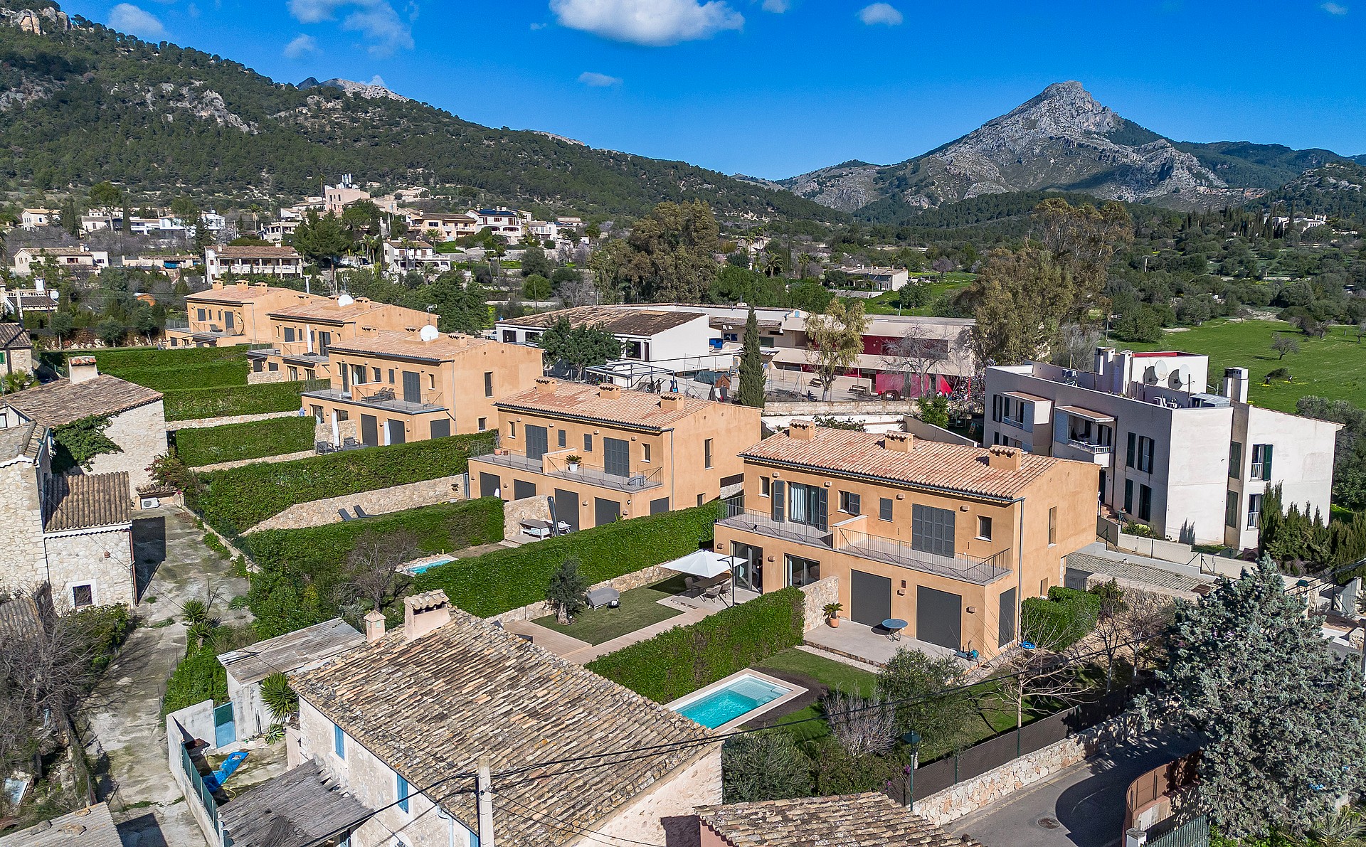 Aerial drone view showing a row of terraced townhouses with ochre facades, landscaped gardens, and a pool in Es Capdella.