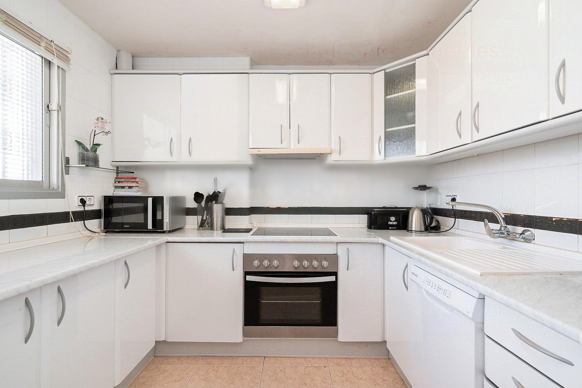 A bright galley kitchen with white cabinets, light countertops, and dark tiled backsplash, illuminated by natural light.
