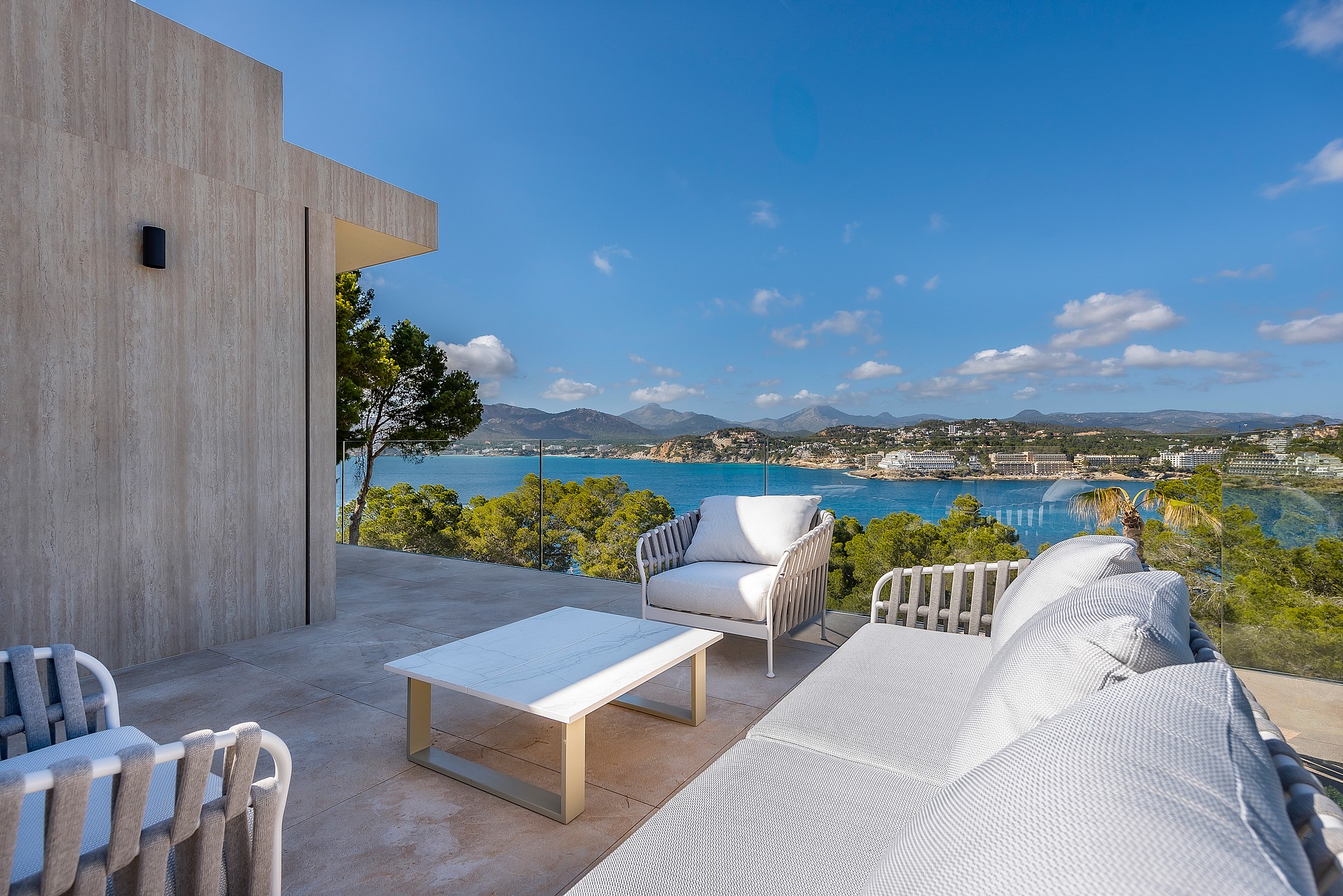 Outdoor terrace with white lounge furniture, glass railings, and a wide view of the Mediterranean bay and coastal mountains.