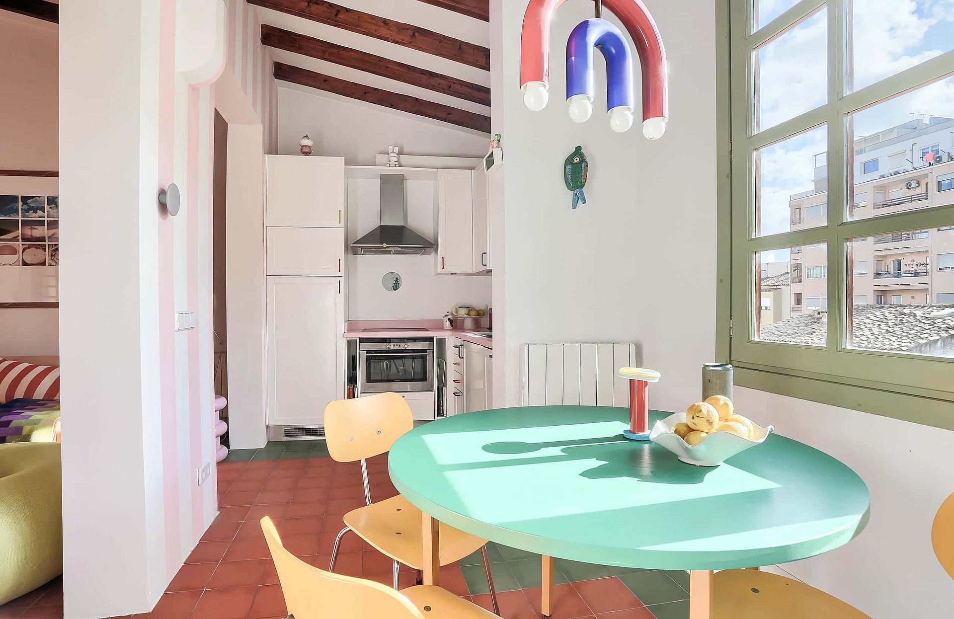A bright kitchen area with a teal round dining table, exposed wooden ceiling beams, white cabinetry, and large windows overlooking the city.