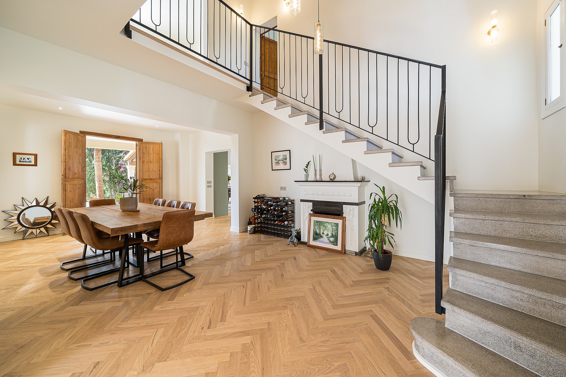 Sunlit living space with oak chevron floors, a black iron staircase, and a dining table near open wooden shutters.
