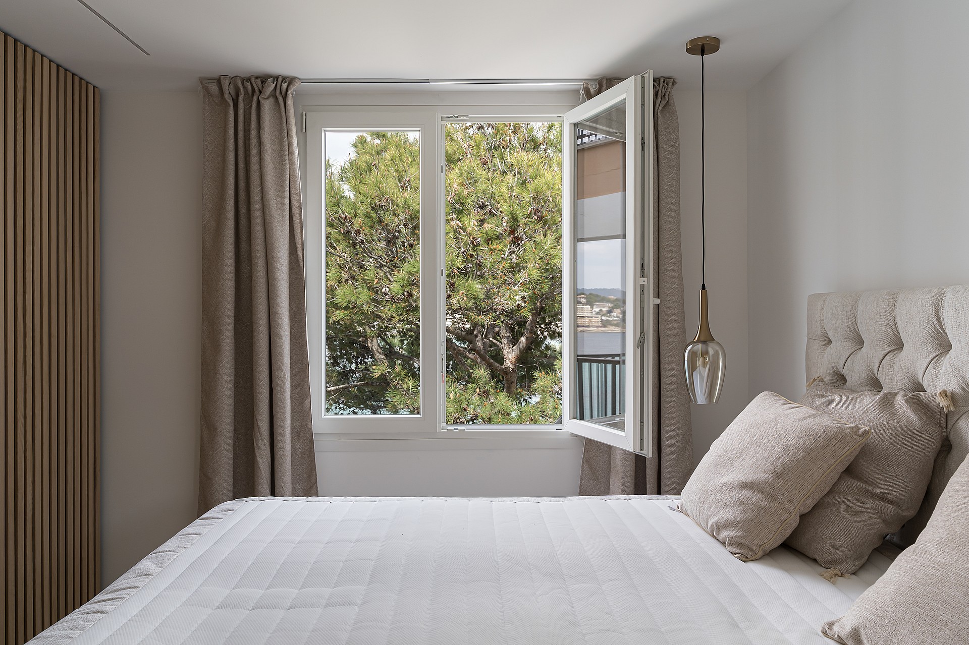A bright bedroom with white linens, neutral curtains, and an open window overlooking pine trees and a distant coastline.